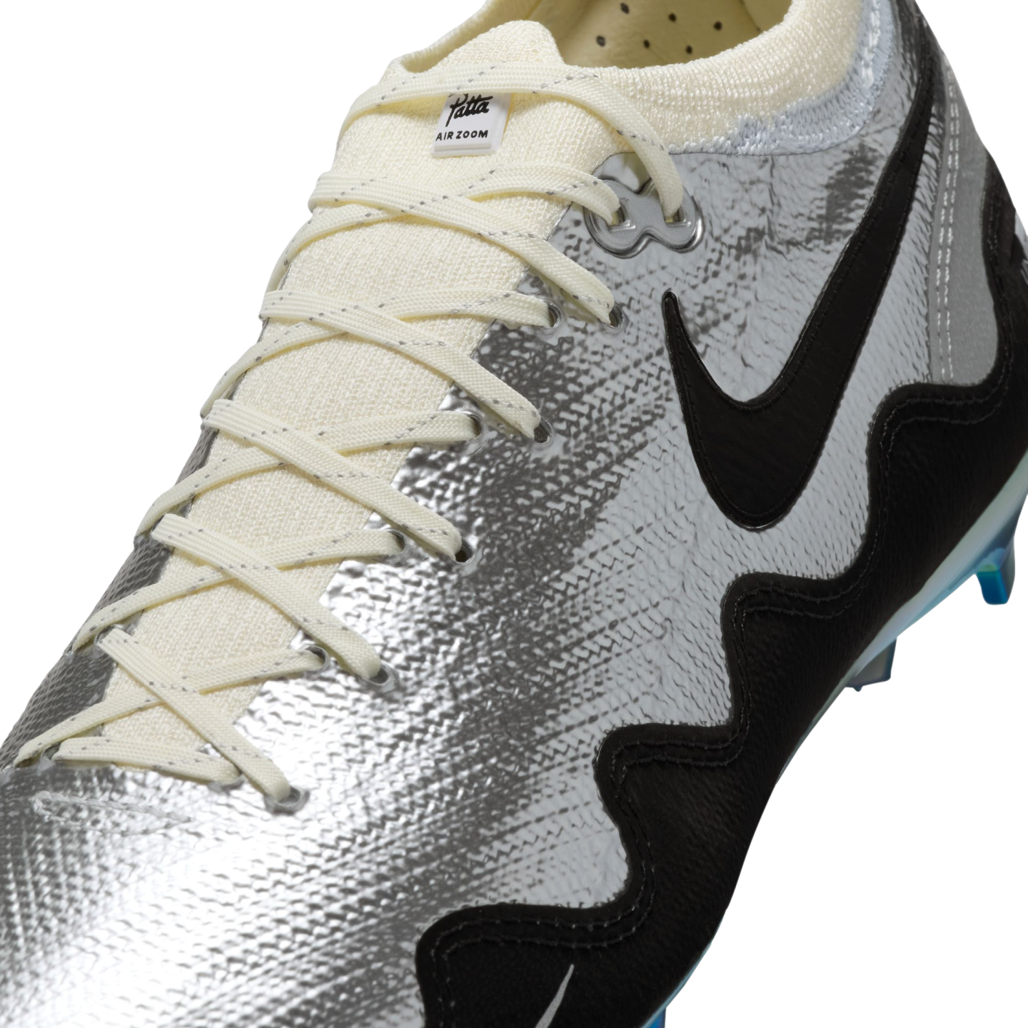 A close-up shot of a Nike Mercurial soccer cleat featuring a textured metallic silver upper with a prominent black swoosh and wavy black graphic along the side. The shoe has off-white laces and a small "Patta AIR ZOOM" label on the tongue, set against a solid black background.