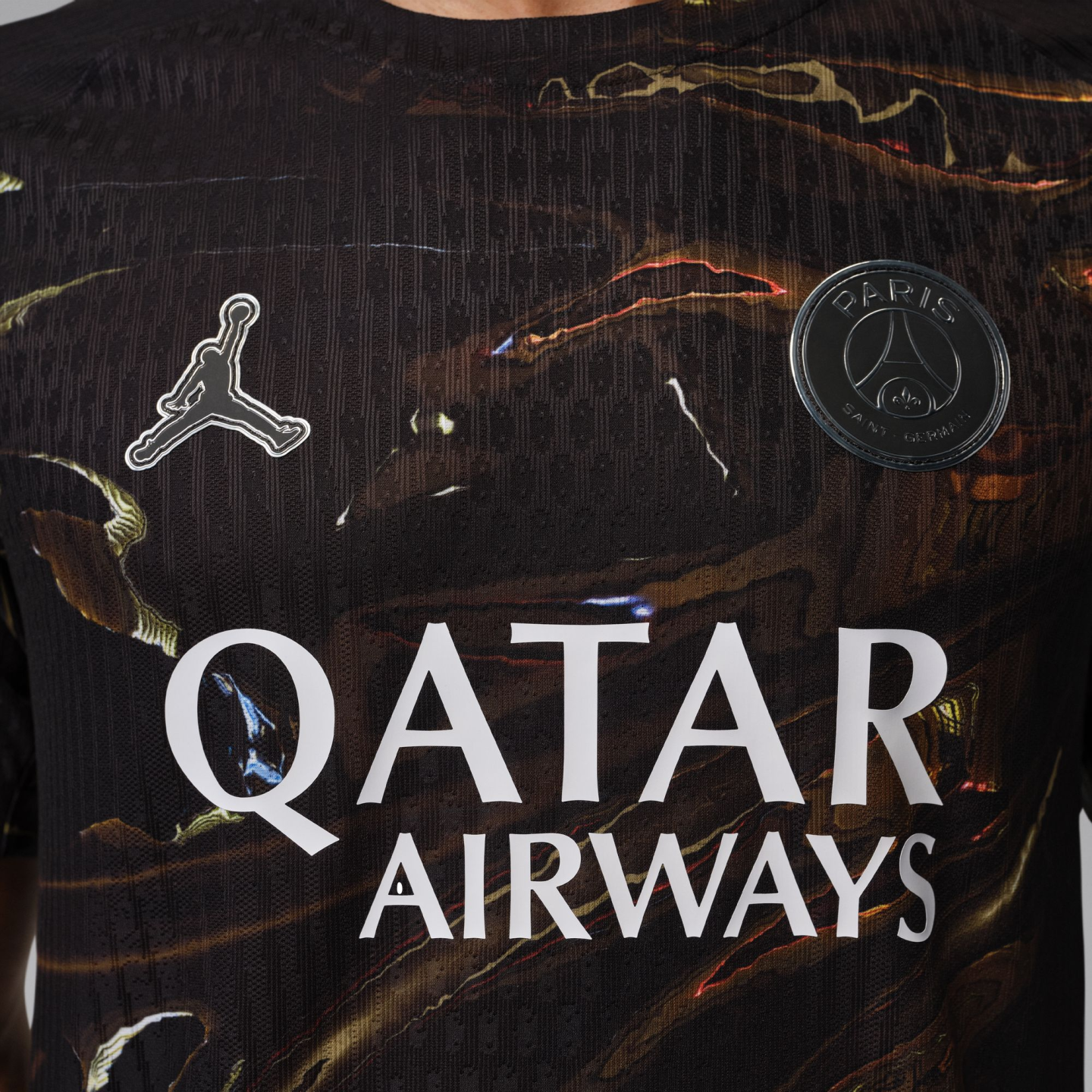 A close-up of a black Paris Saint-Germain soccer jersey with a brown and gold marble-like pattern. It features the white Jordan Brand "Jumpman" logo on the right chest, a black and silver PSG crest on the left chest, and the "QATAR AIRWAYS" sponsor logo in bold white text across the center.