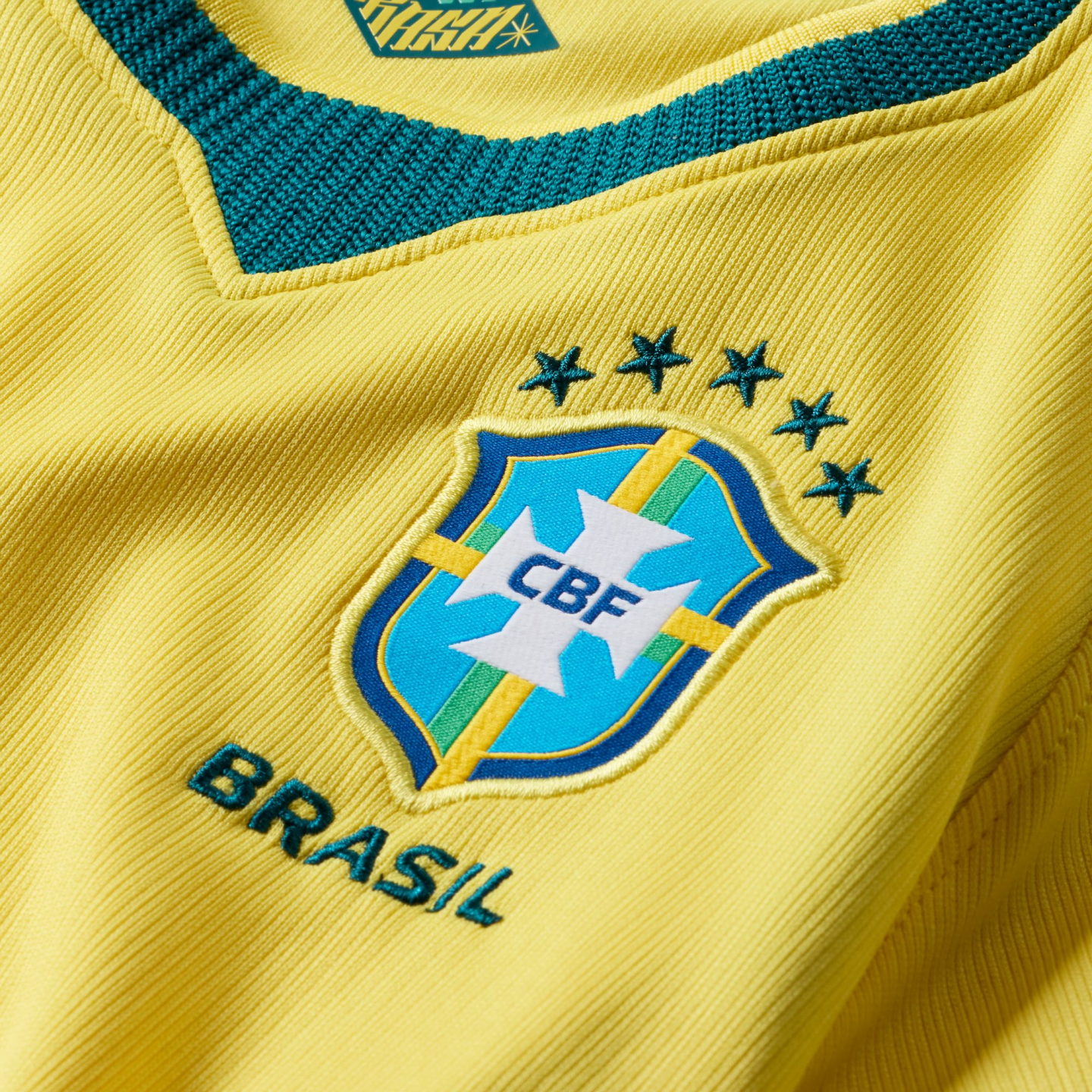 A close-up of the yellow ribbed fabric of a Brazilian national football jersey. The blue and white CBF crest is featured in the center, topped by five green stars and with the word "BRASIL" embroidered in green below it. A dark green, textured V-neck collar is visible at the top.