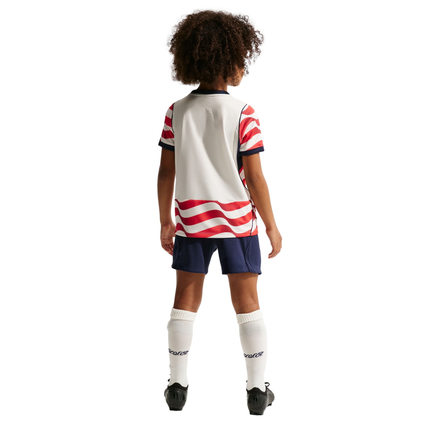 Back view of a child with curly hair wearing a white and red wavy-striped soccer uniform, blue shorts, and white socks against a black background.