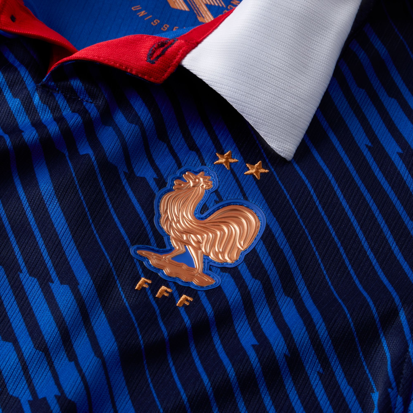 Close-up of a blue France national football jersey featuring a gold Gallic rooster crest, two stars, and 'FFF' lettering over a blue striped pattern with a white collar.