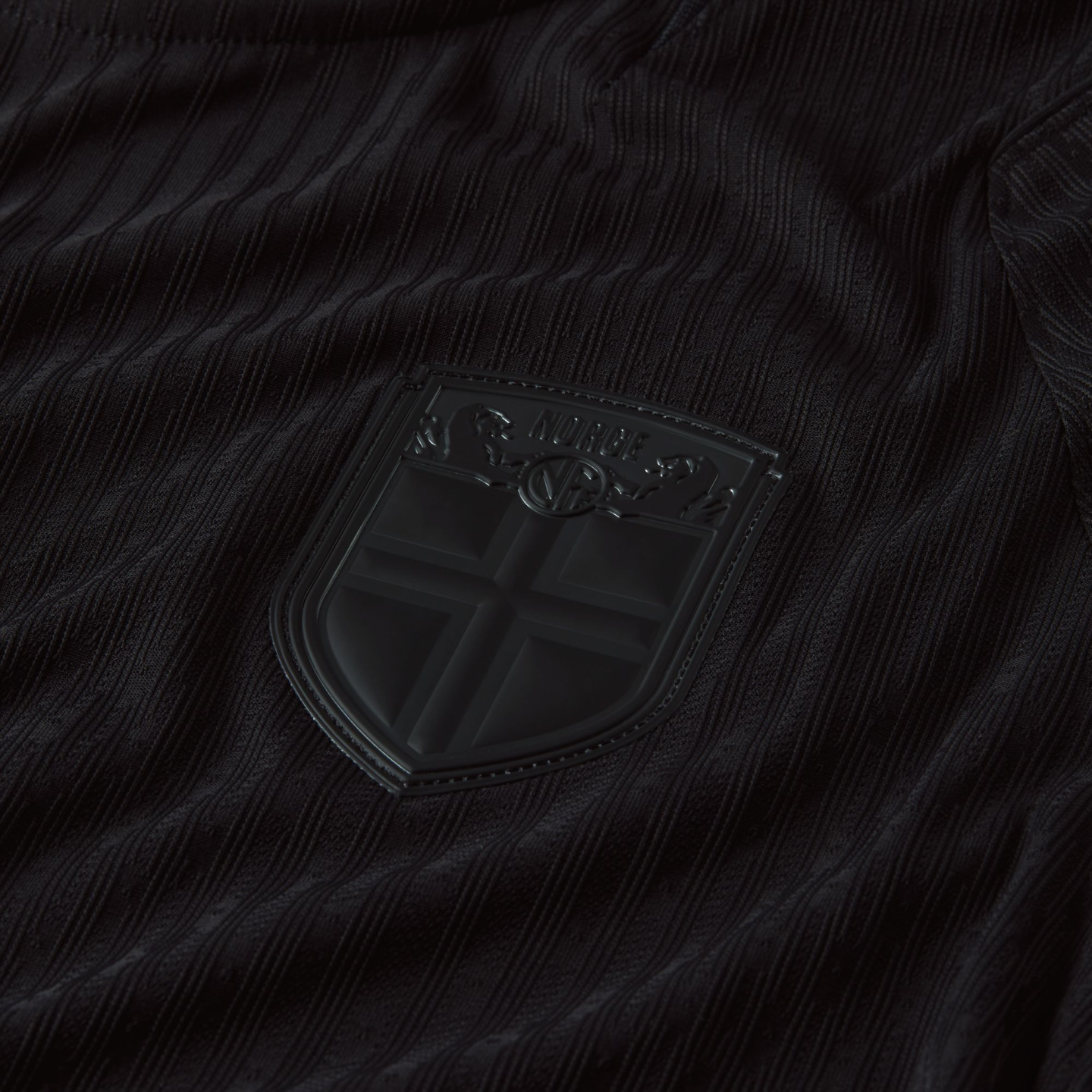 Close-up of a black textured jersey featuring a matte black shield crest with the word "NORGE" and a Nordic cross motif.
