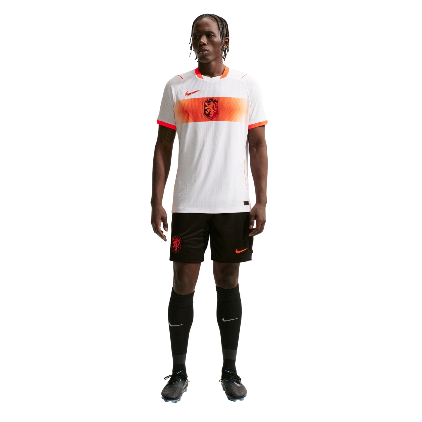 A full-length portrait of a male athlete wearing a Netherlands national soccer team away kit, standing against a solid black background. He is wearing a white jersey with an orange horizontal stripe across the chest, black shorts, and black knee-high socks.