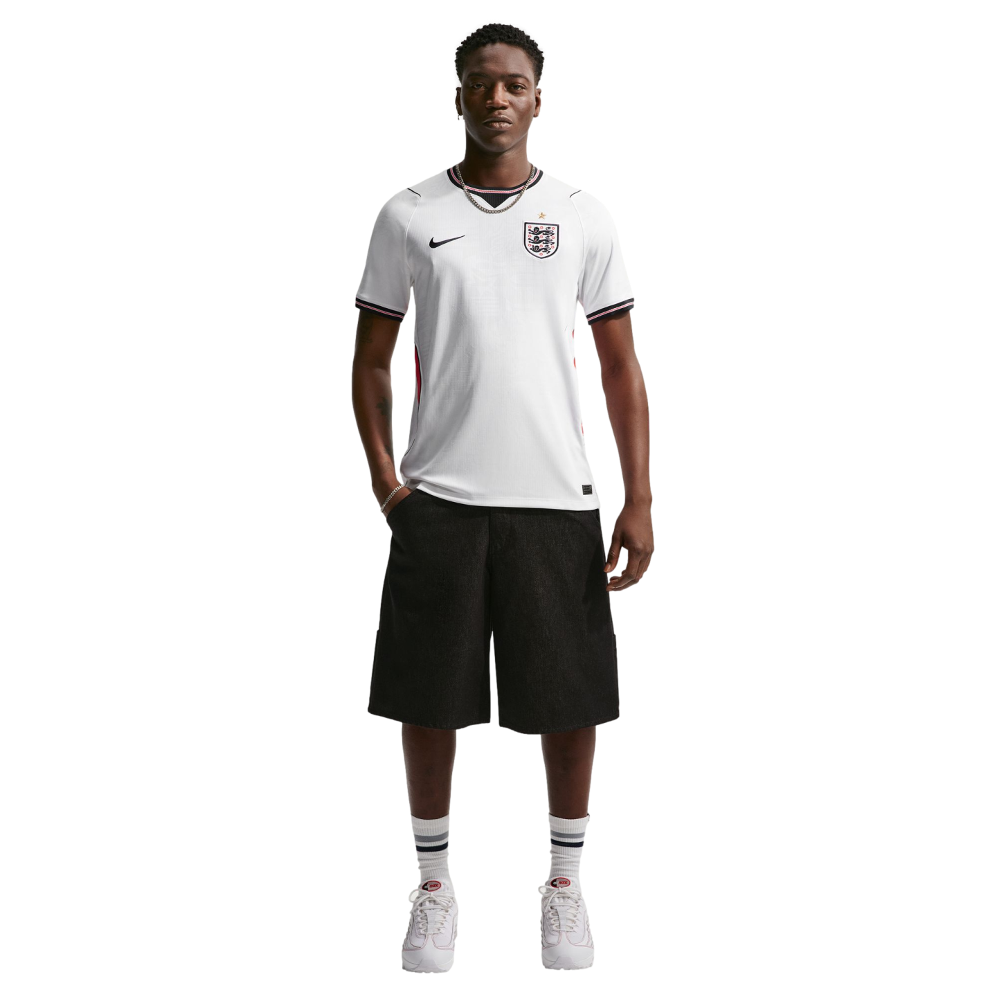 A man stands against a black background wearing a white England soccer jersey, black shorts, and white sneakers.