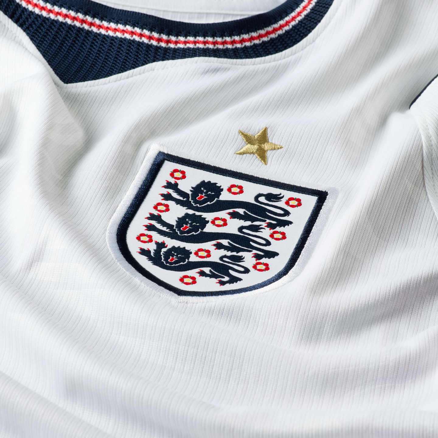 Close-up of a white England national football jersey showing the Three Lions crest with a gold star above it on ribbed fabric, and part of a navy, red, and white striped collar.