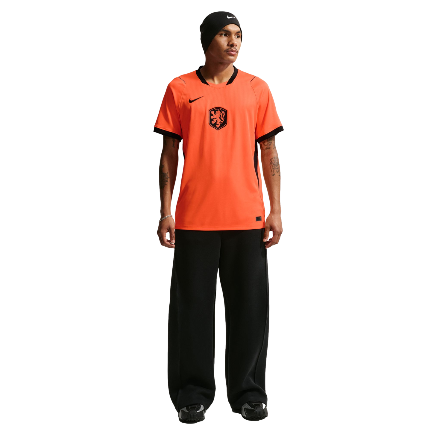 A studio photograph of a man standing against a black background. He is wearing a bright orange Netherlands national football team jersey with black trim, wide-leg black trousers, and a black beanie.