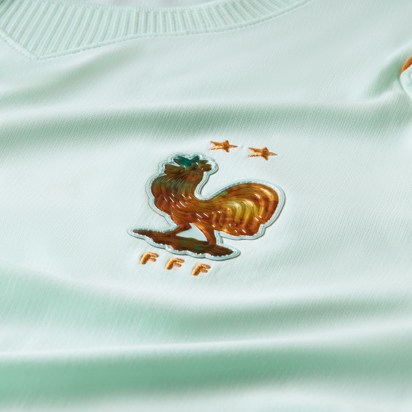 Close-up of a mint green soccer jersey featuring an iridescent French Football Federation (FFF) rooster crest with two gold stars above it.