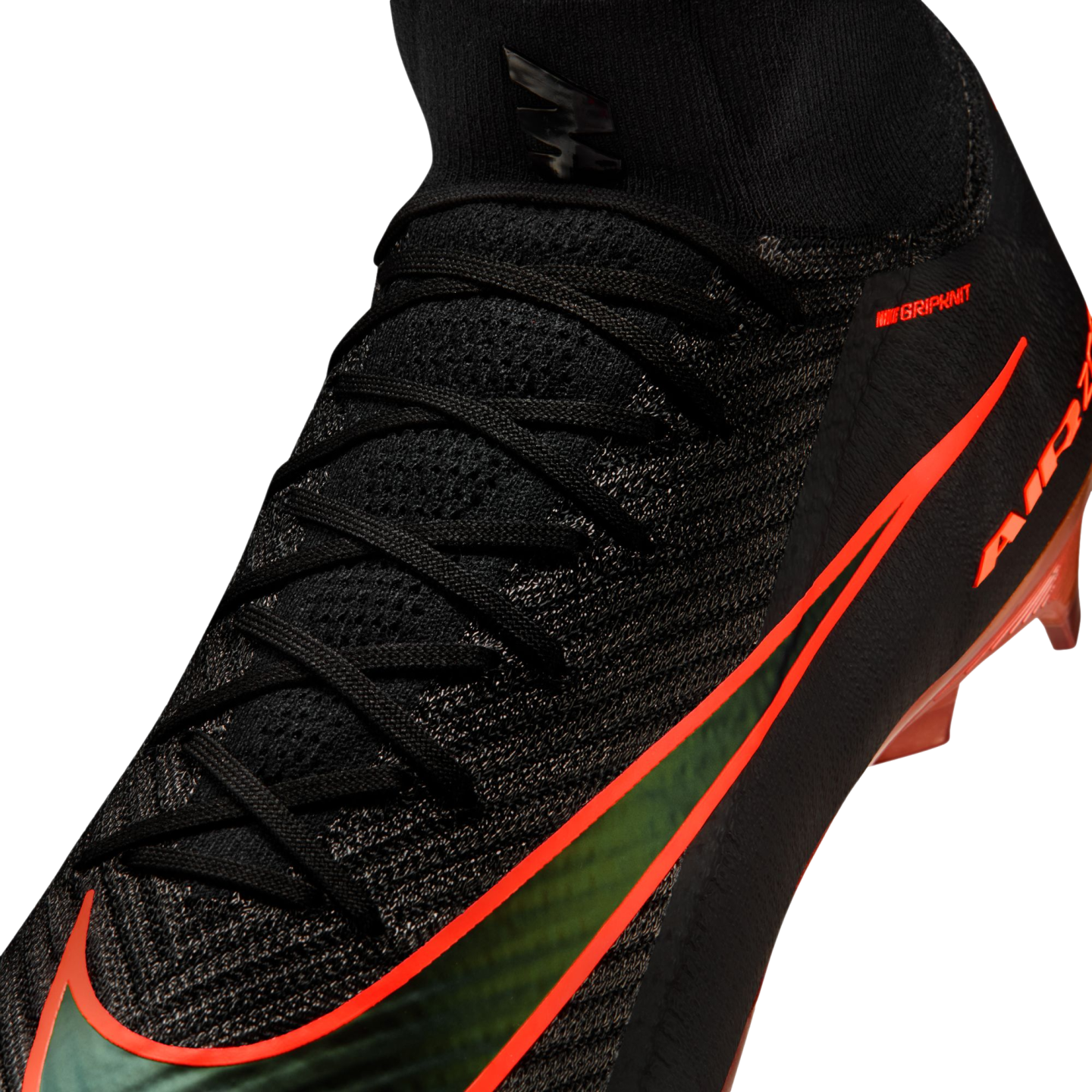 A close-up view of a black Nike soccer cleat with a textured knit upper and a sock-like collar. The shoe features a large, iridescent green Nike swoosh outlined in bright orange, along with small orange "NIKE GRIPKNIT" text and orange studs visible at the base.