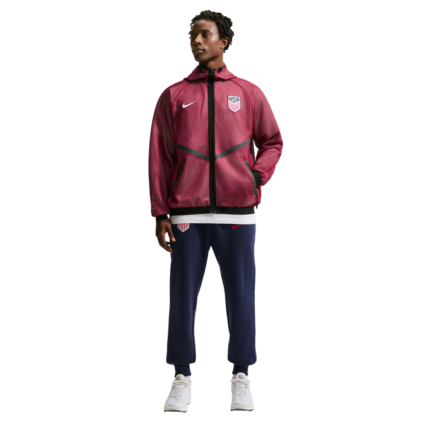 A full-length photo of a man modeling Nike USA soccer apparel against a black background. He wears a maroon hooded jacket with a white swoosh and USA crest, navy joggers with matching logos, and white sneakers.