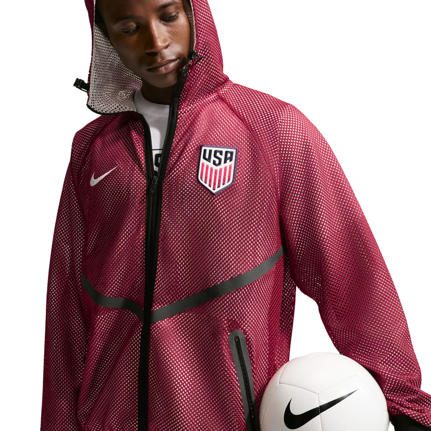 A young Black man looking down while wearing a maroon mesh hooded jacket featuring a "USA" soccer crest and a white Nike swoosh. He holds a white Nike soccer ball against his hip, set against a solid black background.
