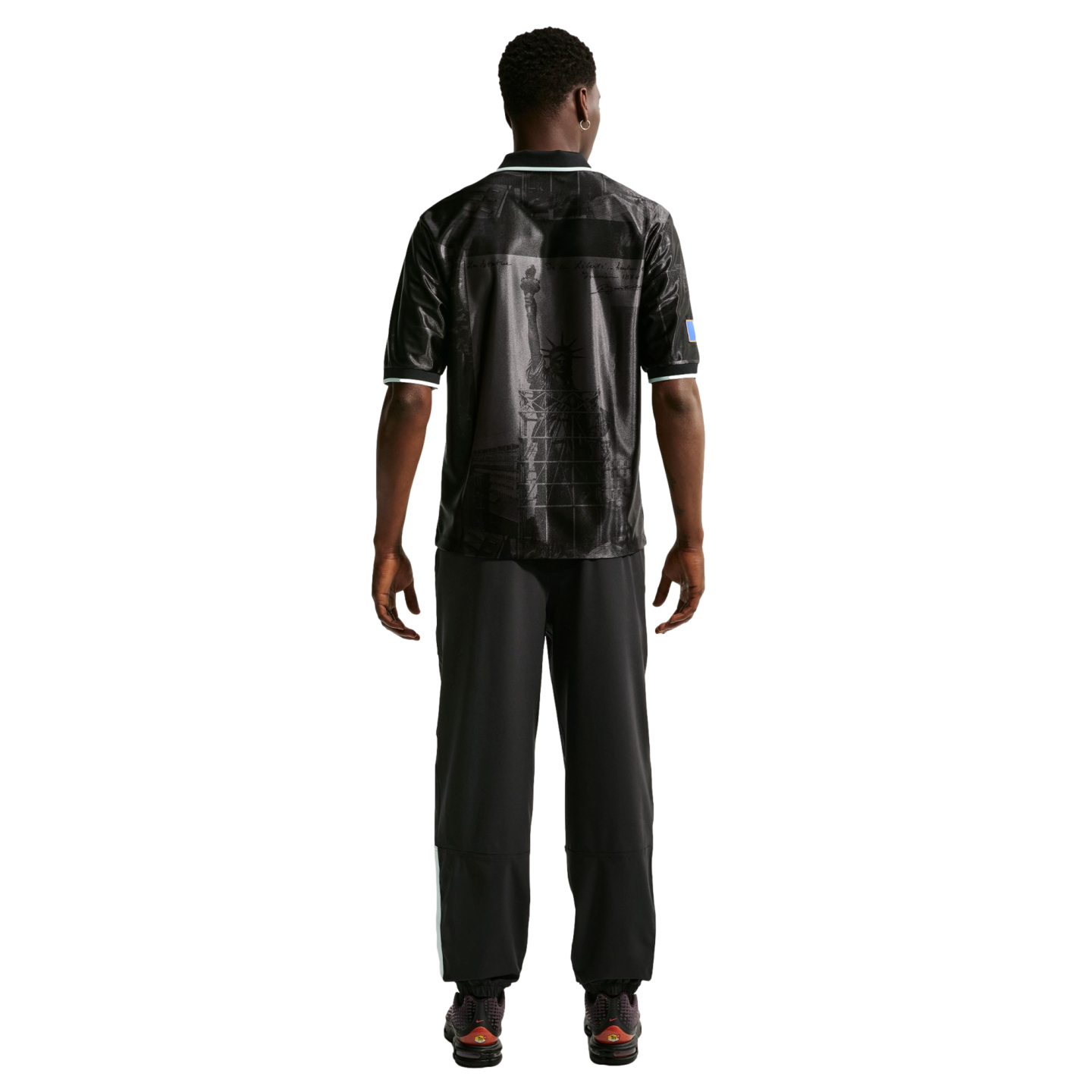 Rear view of a man in a black polo shirt with a Statue of Liberty graphic, black pants, and dark sneakers against a white background.