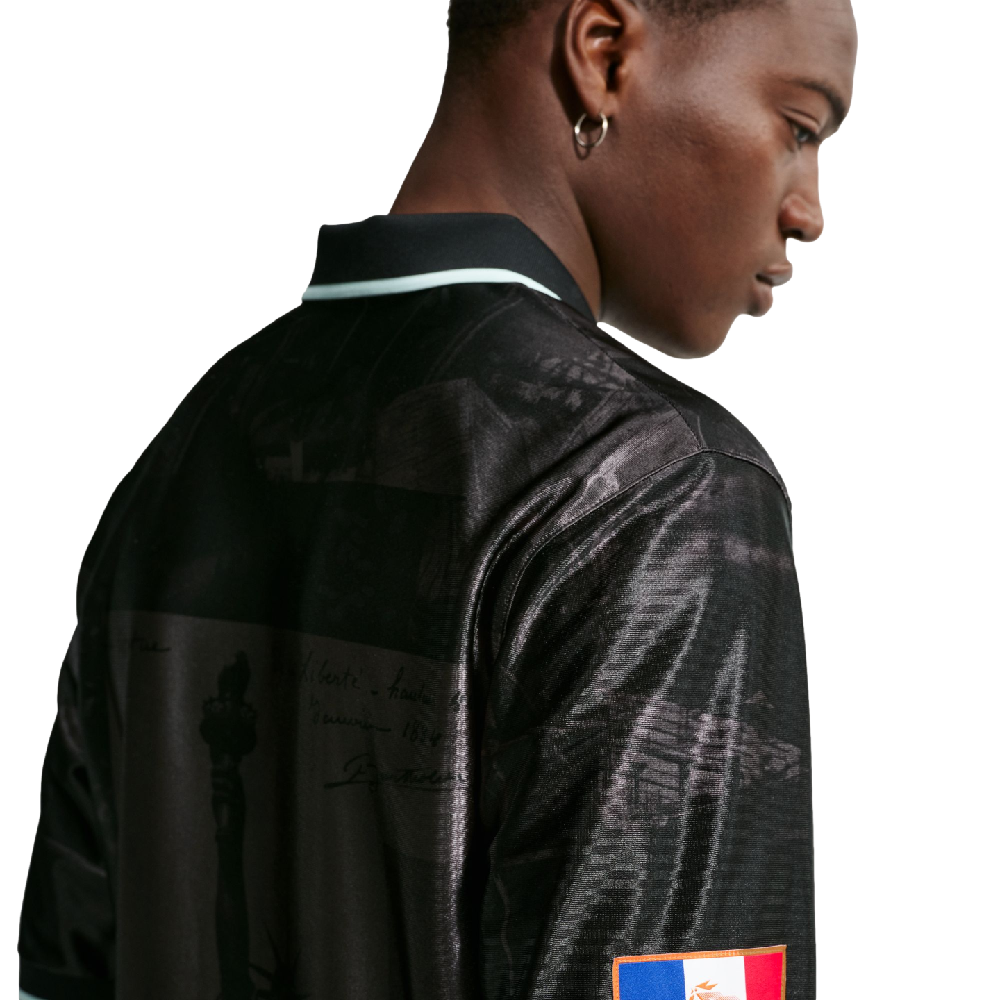 Profile of a Black man from behind, wearing a shiny black polo shirt with historical graphic prints and a small flag patch on the sleeve.