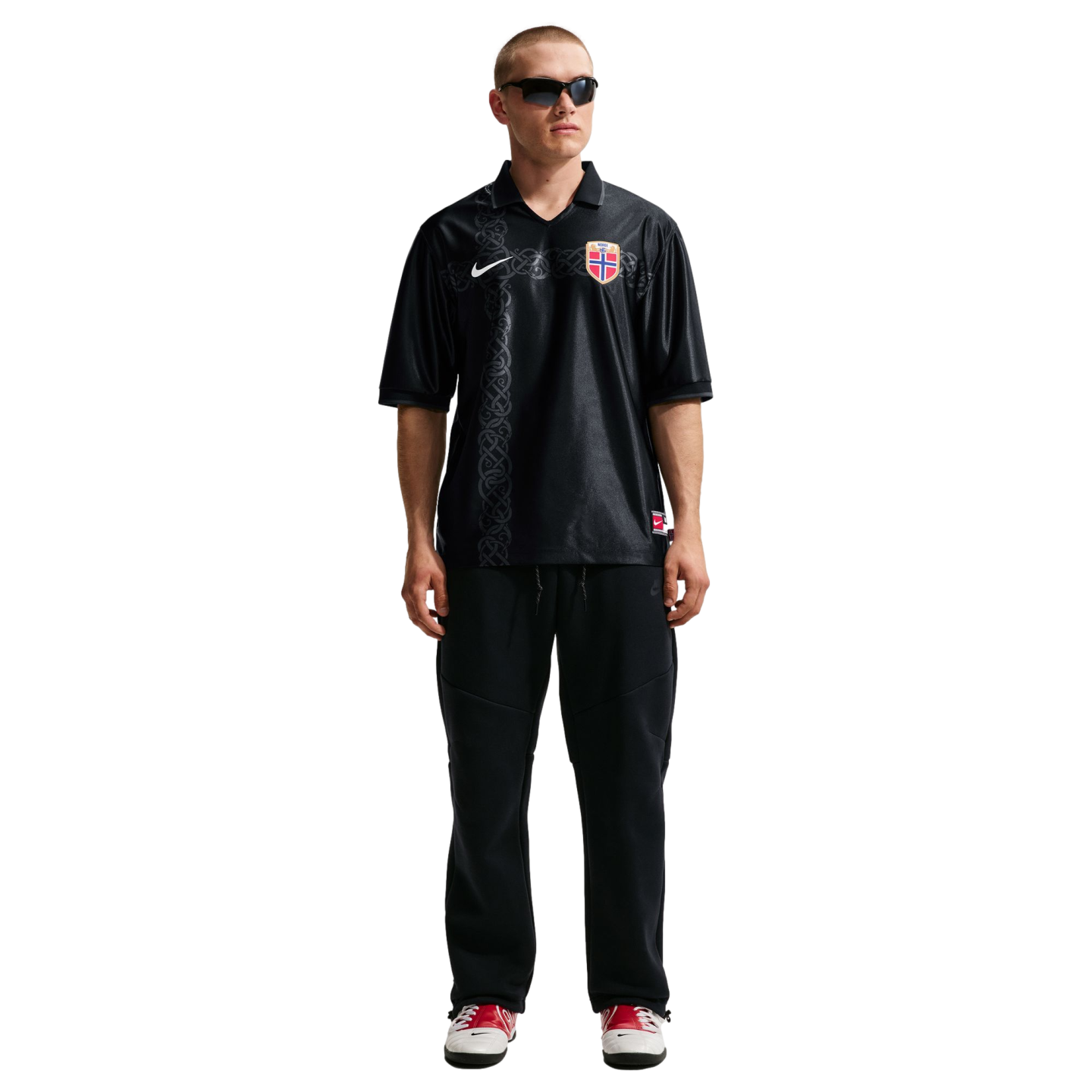 A man modeling a black Nike Norway football jersey with a Nordic knot pattern, black sweatpants, sunglasses, and red and white sneakers against a white background.