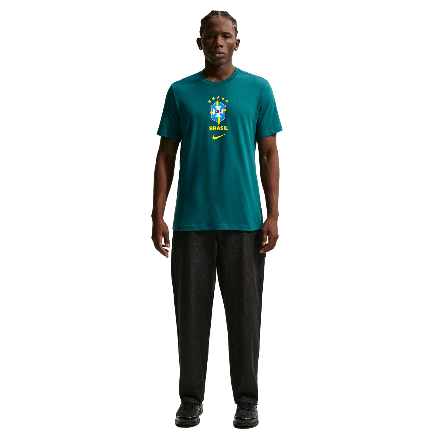 A young man stands centered against a solid black background, wearing a teal green Nike t-shirt featuring the Brazilian football federation (CBF) crest. He is also wearing dark charcoal straight-leg trousers and black sneakers.