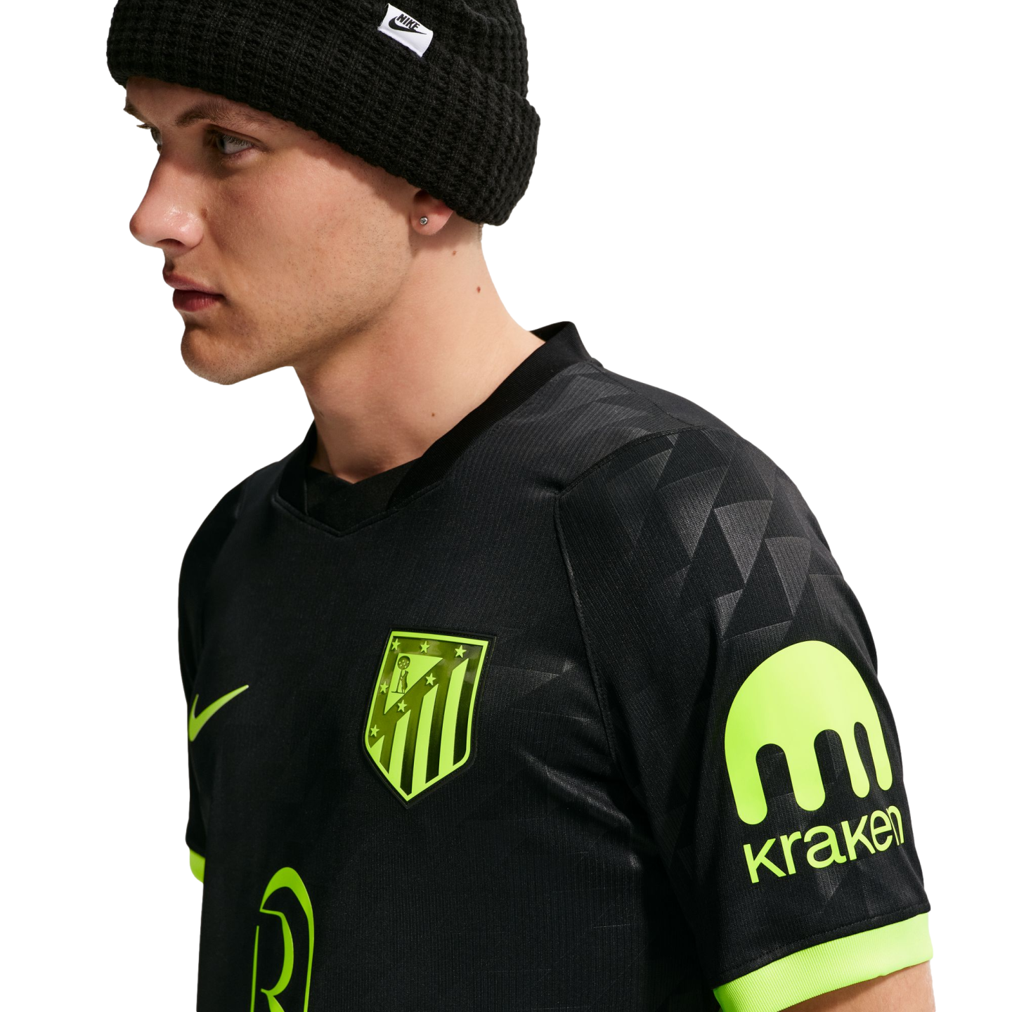 A man in profile wearing a black Nike beanie and a black Atletico Madrid jersey with neon green logos and a Kraken sponsor patch on the sleeve.