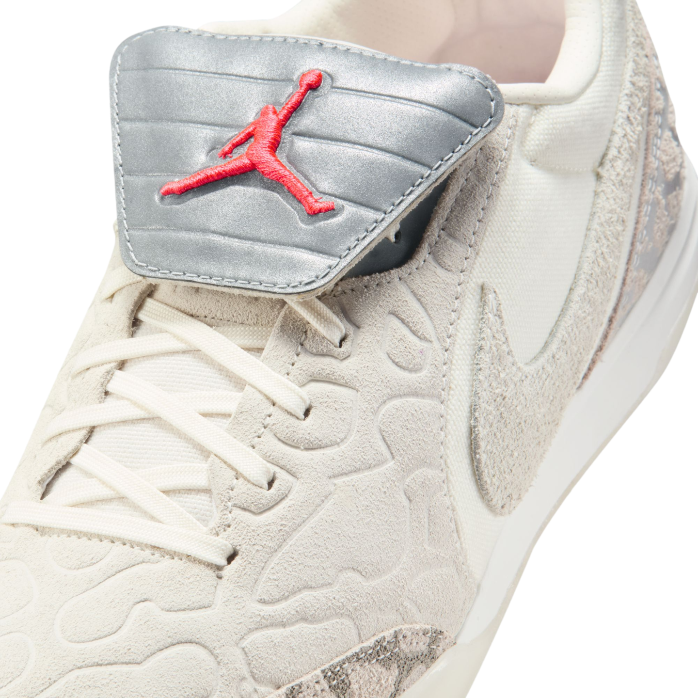 Close-up of a beige Jordan sneaker with textured overlays, white laces, and a metallic silver tongue flap featuring a red embroidered Jumpman logo.