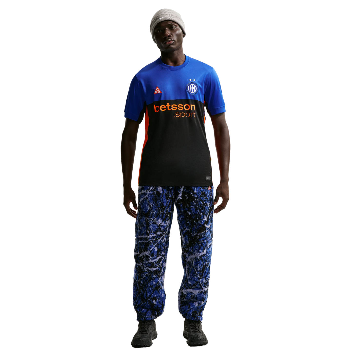 A man models a blue and black Inter Milan x Nike ACG jersey, blue patterned trousers, a grey beanie, and dark sneakers against a black background.
