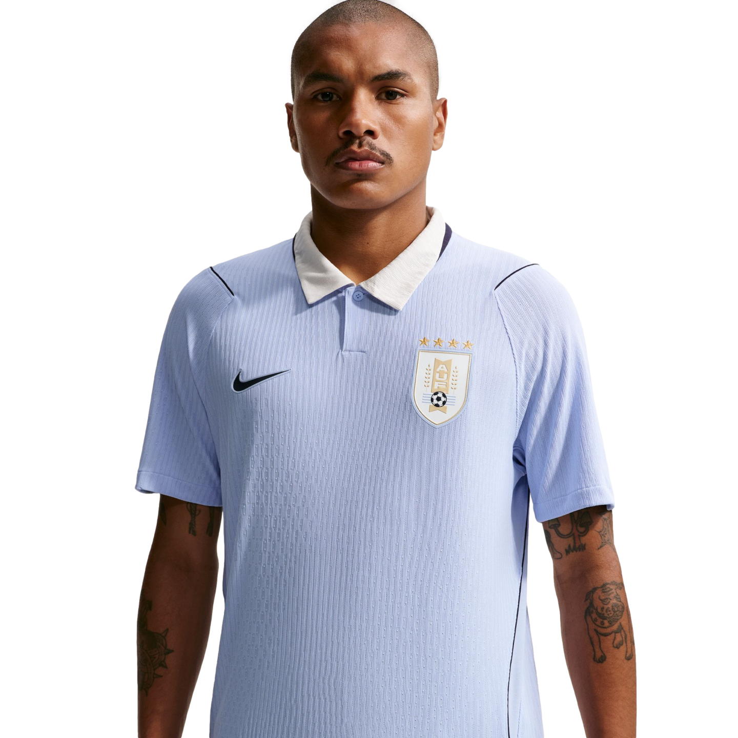 A man with a mustache and tattooed forearms wears a light blue Uruguay national team soccer jersey with a white collar against a solid black background.