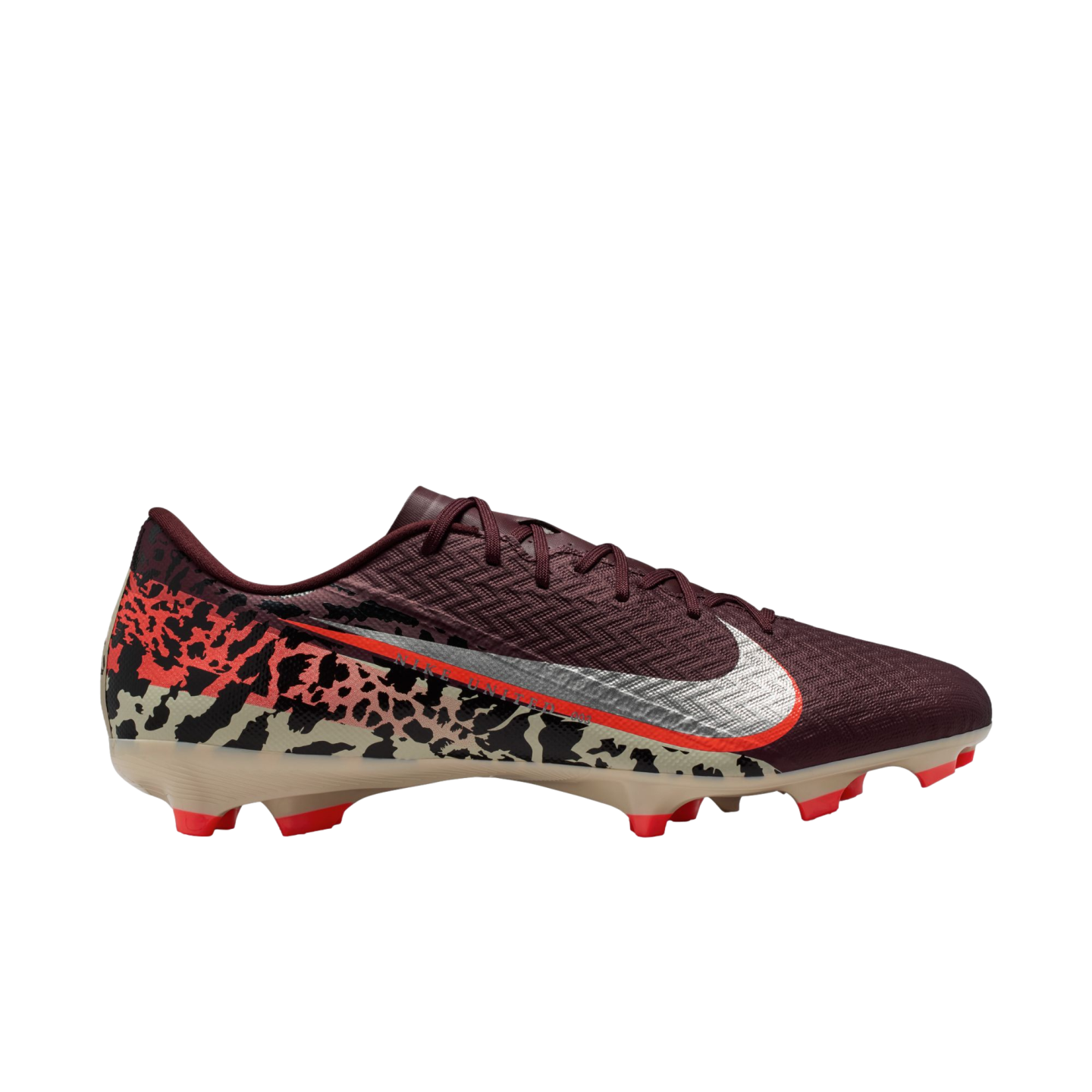 A profile view of a Nike soccer cleat against a solid black background. The shoe features a textured maroon upper, a large silver Nike Swoosh with an orange border, and a bold abstract graphic pattern in tan, black, and orange across the lower half and heel. It has a tan soleplate with bright orange studs.