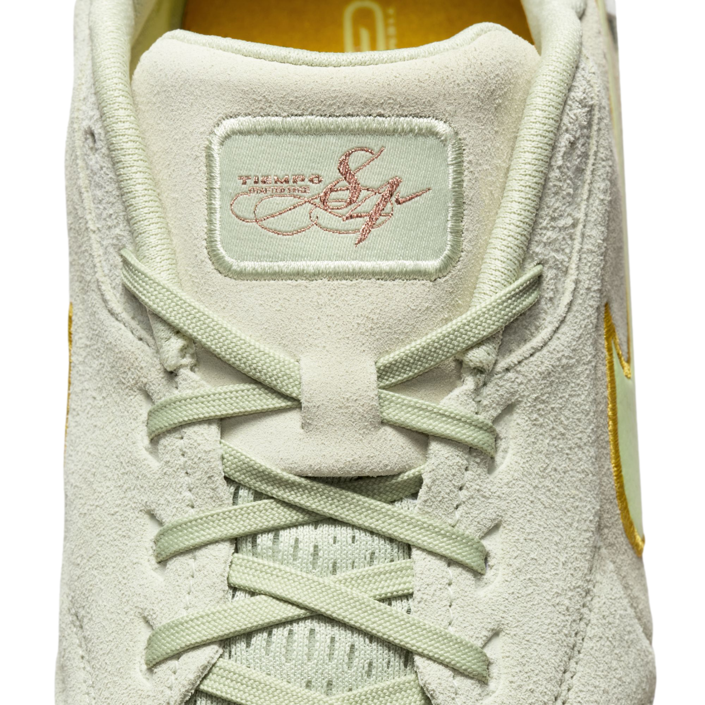 Close-up of a light green suede Nike Tiempo shoe's tongue and laces. A rectangular patch features "TIEMPO" and a rose-gold embroidered signature.