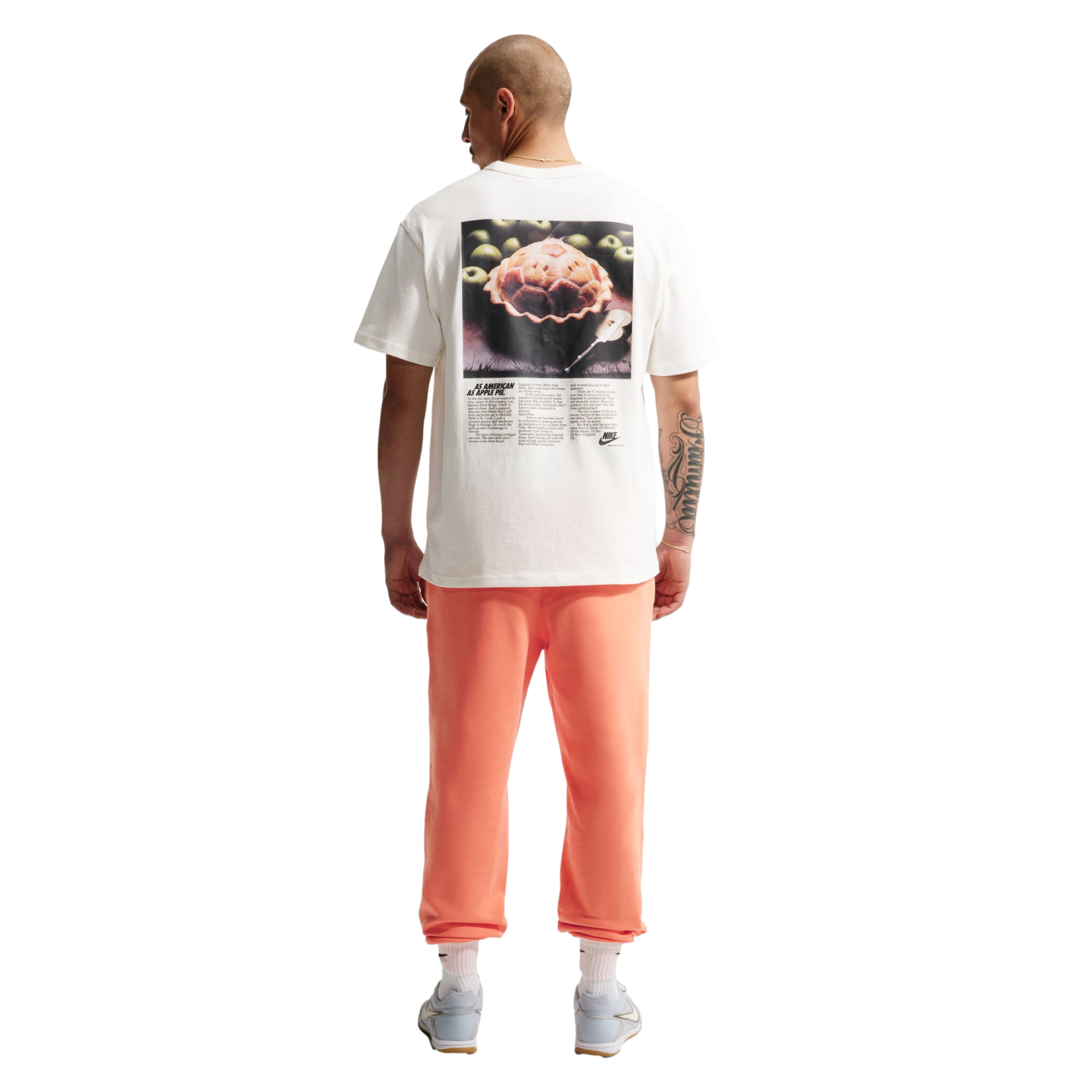 A back view of a man standing against a black background, wearing a white t-shirt with a large graphic of an apple pie and the text "AS AMERICAN AS APPLE PIE." He is also wearing coral-colored sweatpants, white socks, and light blue sneakers. He has a bald head and a large tattoo on his right forearm.