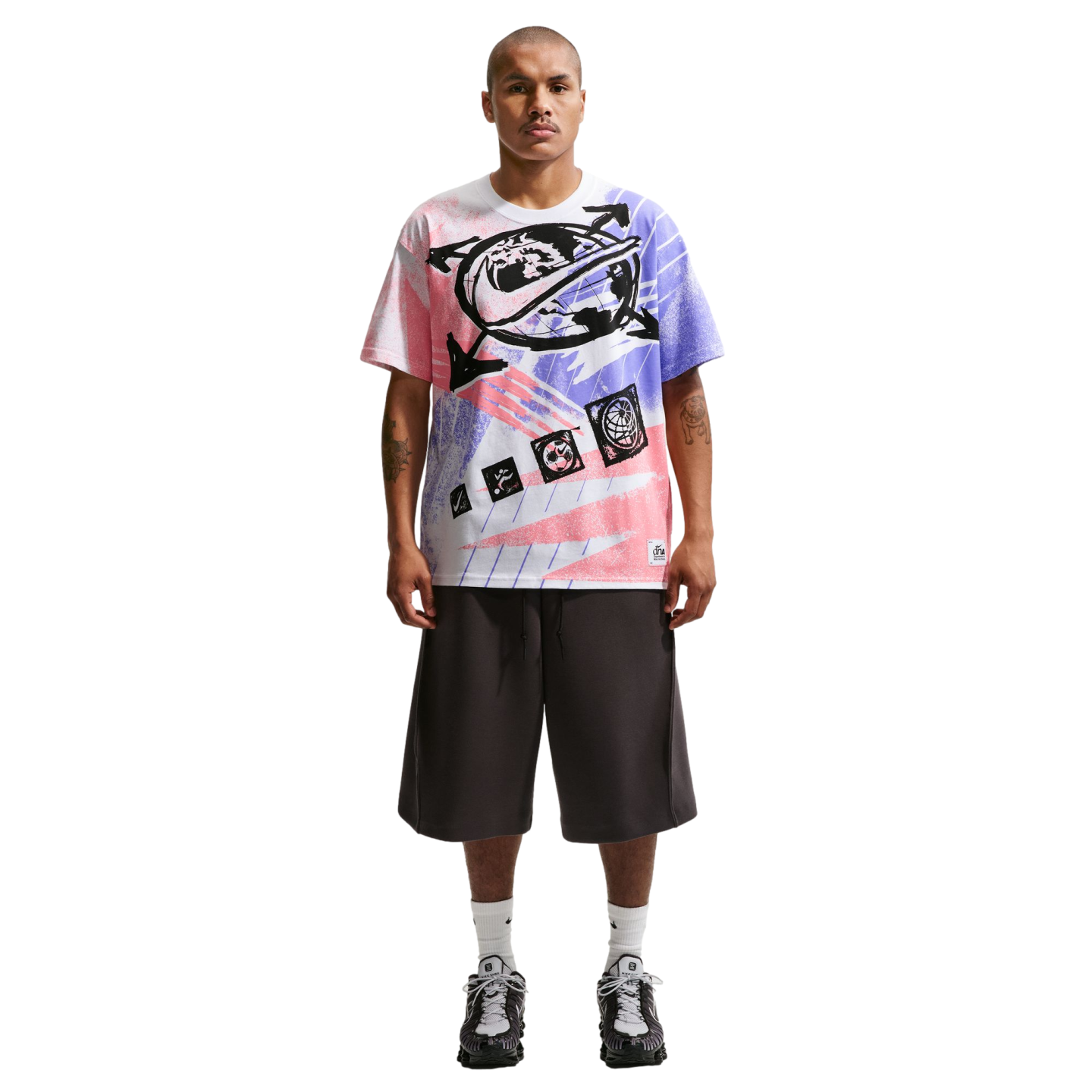 A full-length photo of a man standing against a black background, wearing an oversized graphic t-shirt with pink and purple spray-paint designs, wide-leg charcoal grey shorts, white crew socks, and grey athletic sneakers. He has short hair, a mustache, and tattoos on both forearms.