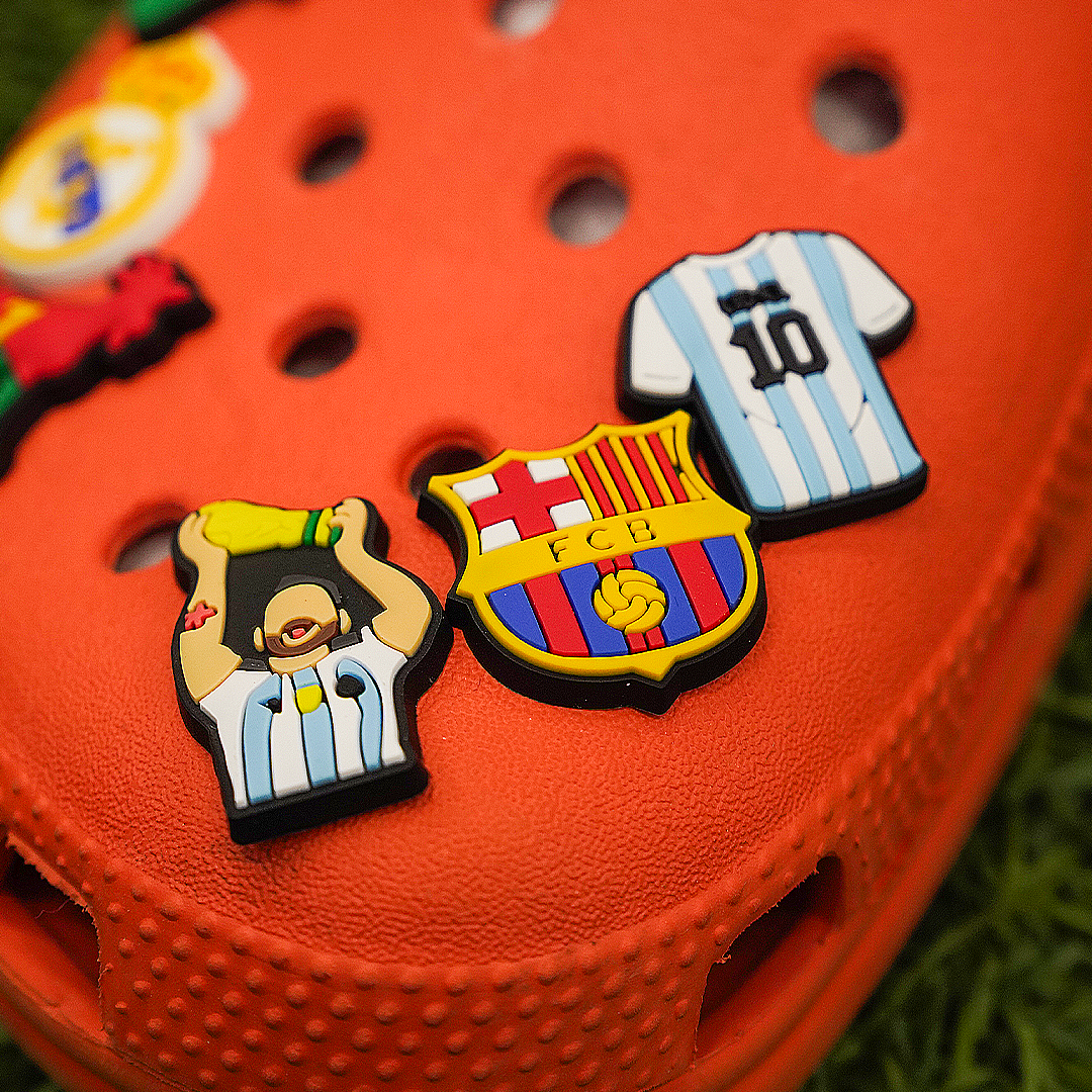 A close-up of an orange shoe featuring several soccer croc charms, including Lionel Messi holding the World Cup trophy, the FC Barcelona crest, and an Argentina national team jersey.