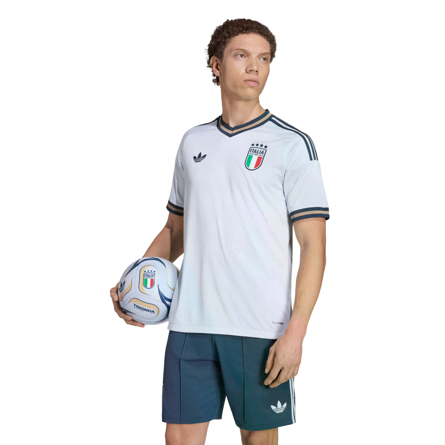 A man poses in a white Italy soccer jersey and blue shorts, holding a football against a black background.