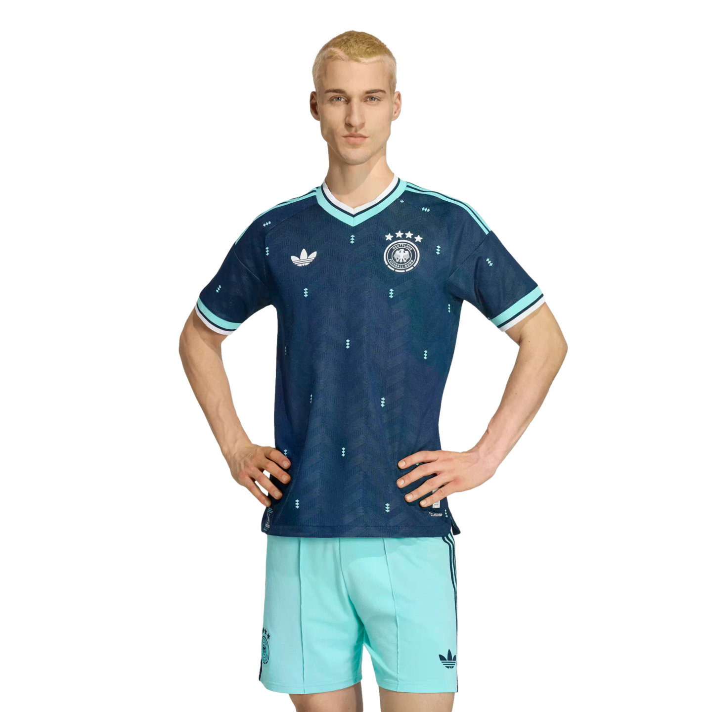 A man models a navy blue and teal German national team soccer kit, standing with hands on his hips against a solid black background.