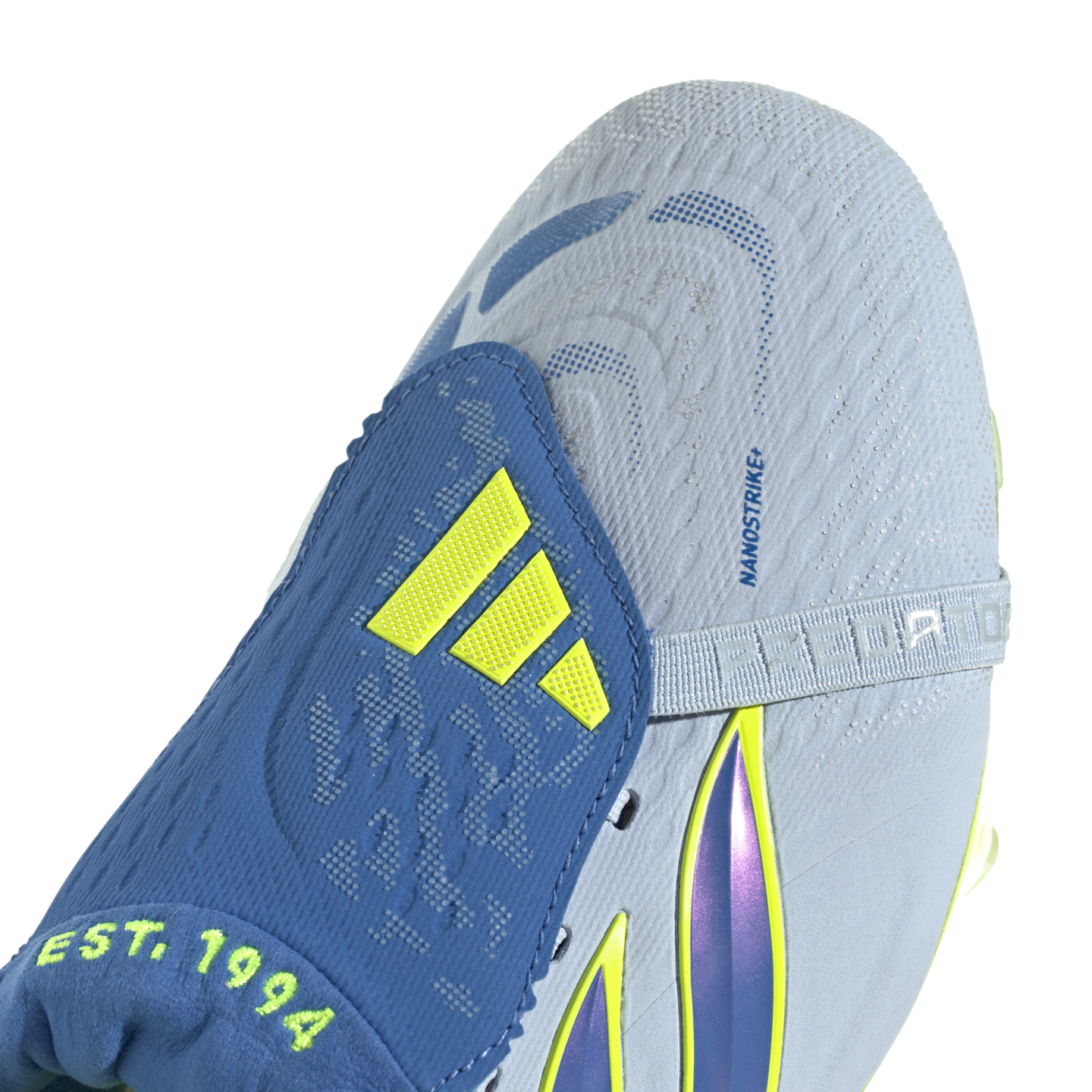 A close-up view of a light blue and blue Adidas Predator soccer cleat. The shoe features a blue fold-over tongue with neon yellow Adidas stripes and the text "EST. 1994" at its base. The light blue textured upper includes the text "NANOSTRIKE+" and a strap embossed with "PREDATOR" across the midfoot. Iridescent purple and neon yellow accents are visible along the side.