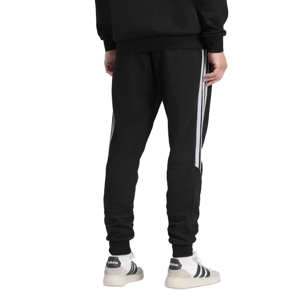 Back view of a person wearing black Adidas track pants with white side stripes and white sneakers.
