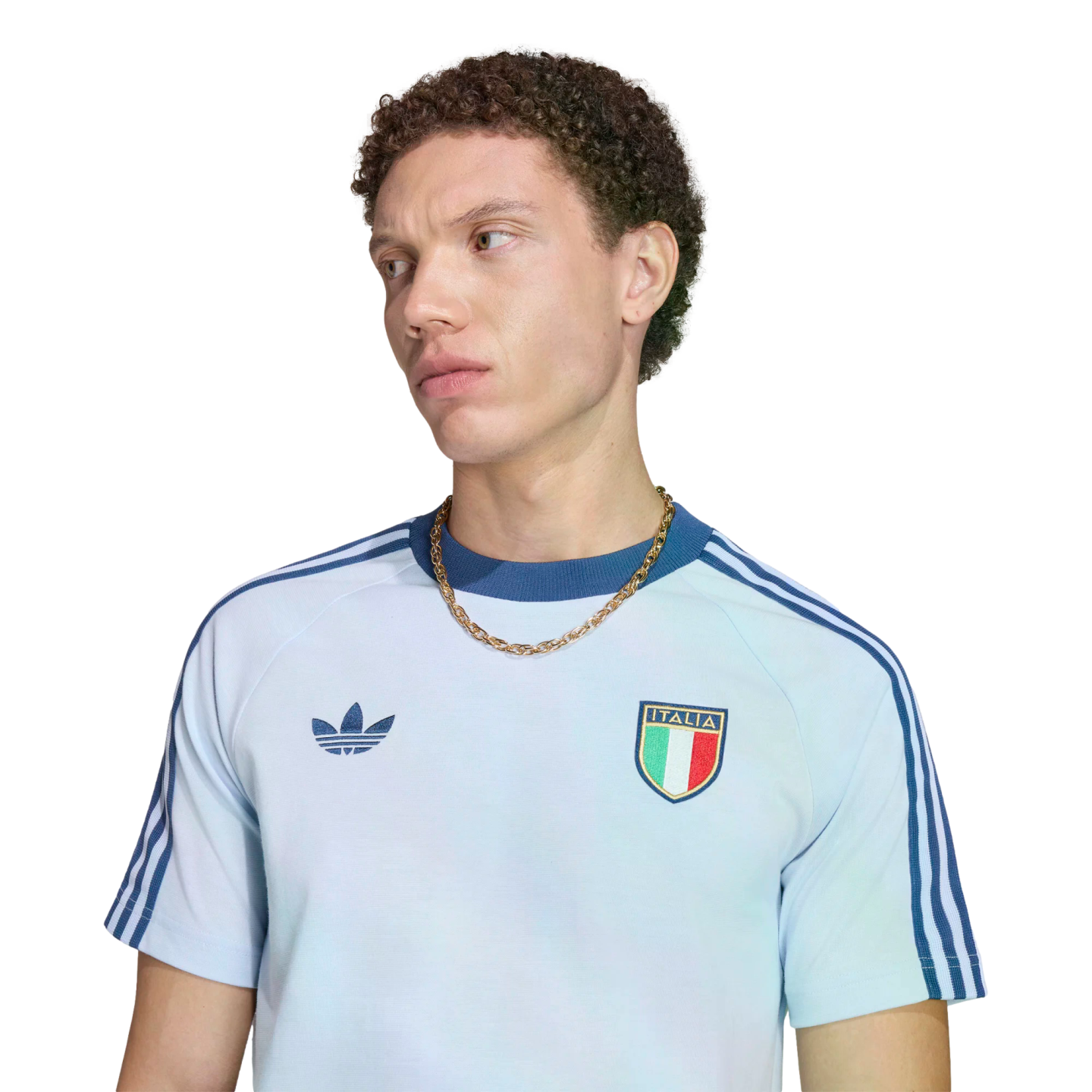 A portrait of a young man with curly brown hair looking away against a black background. He wears a light blue Adidas t-shirt featuring dark blue shoulder stripes, the Adidas logo, the Italian national football team crest, and a chunky gold chain.