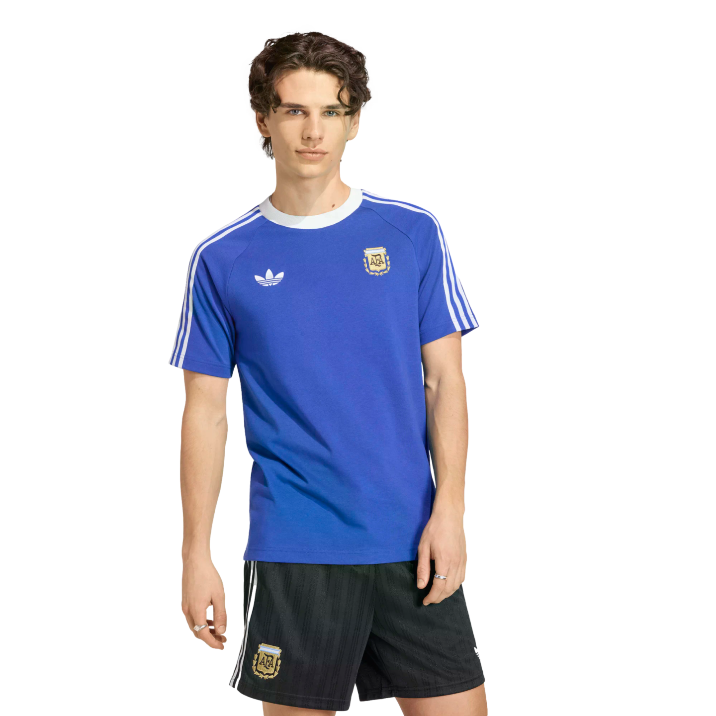 A young man with wavy brown hair models a royal blue Adidas Argentina national football team t-shirt and matching black athletic shorts against a solid black background.