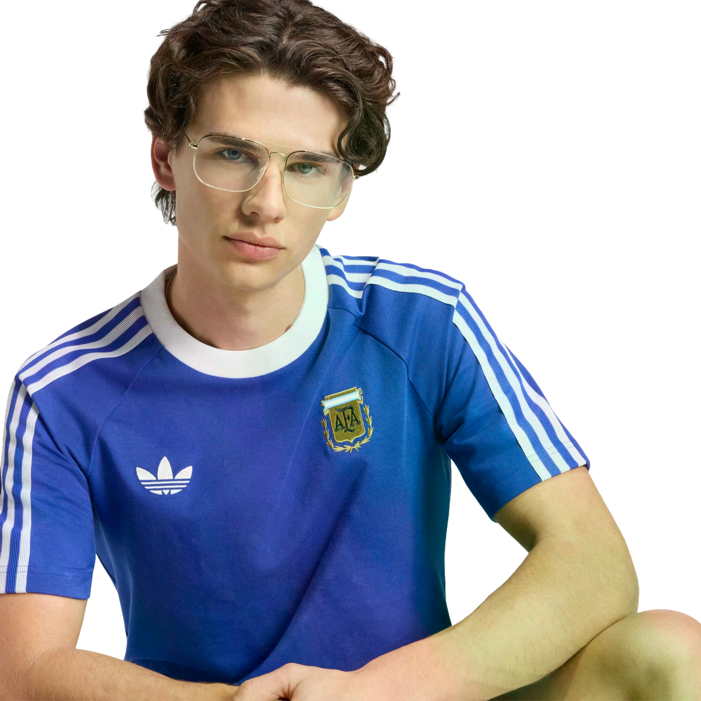 A young man with dark, wavy hair and gold-rimmed glasses wearing a blue Adidas Argentina national football team t-shirt against a black background.