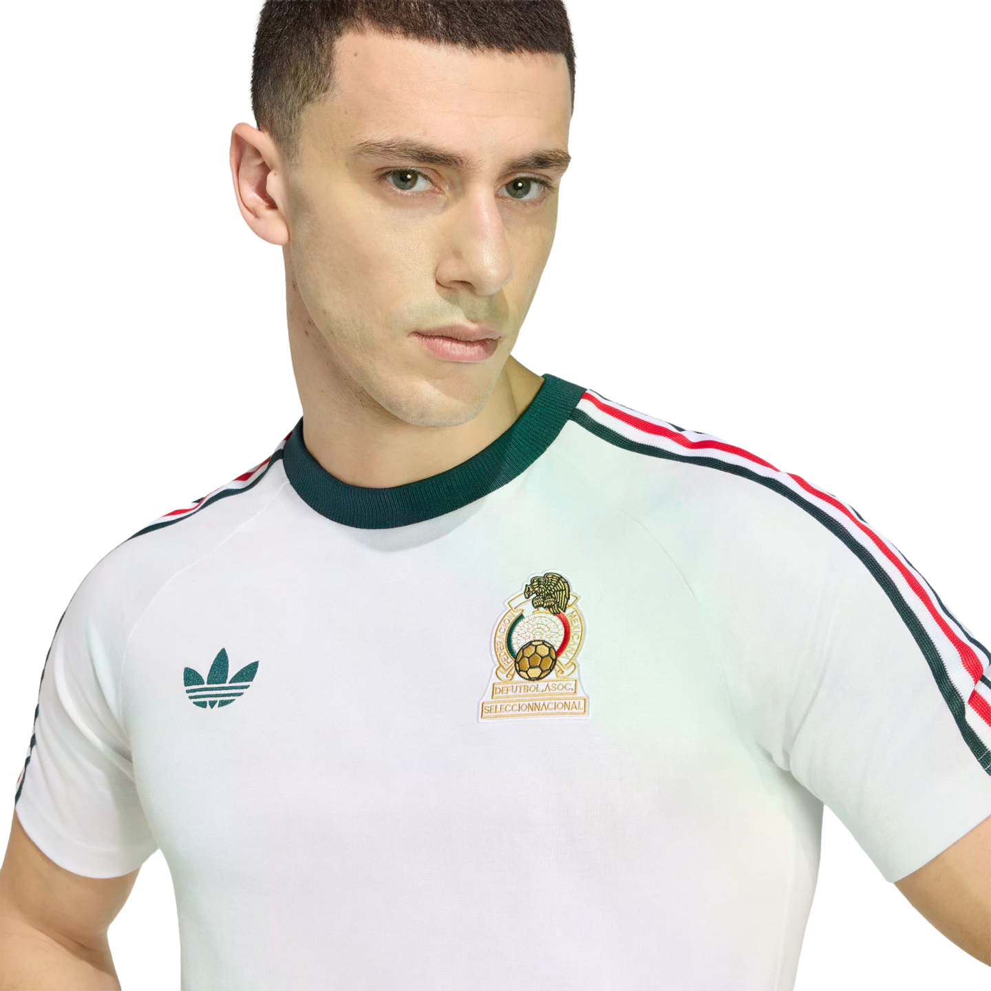 A close-up of a young man wearing a white retro Mexico National Team t-shirt by Adidas. The shirt features a dark green crew neck, a green Adidas Trefoil logo on the right, and the Mexican National Team crest on the left. Three stripes in green, white, and red run down the shoulders against a solid black background.