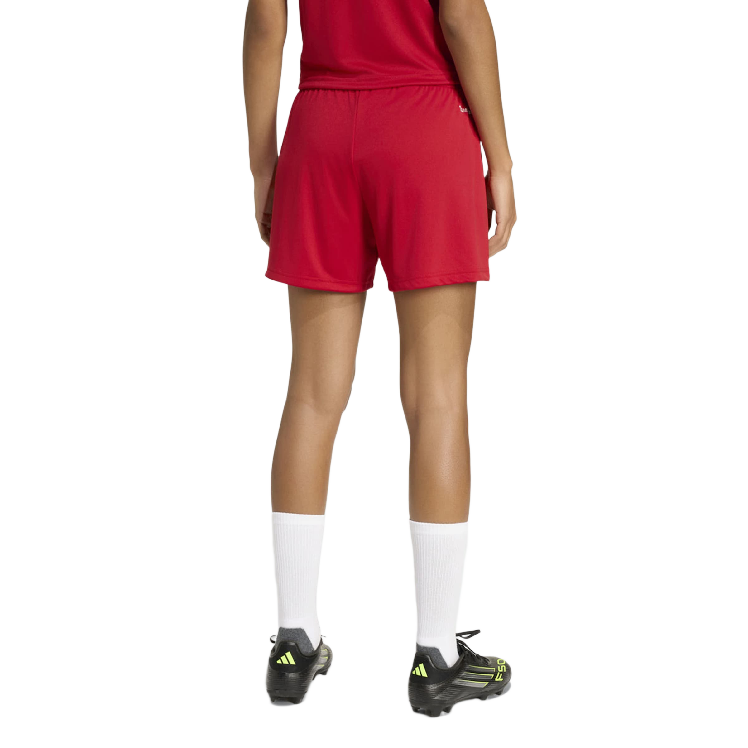Rear view of a person wearing red athletic shorts and shirt, tall white socks, and black Adidas soccer cleats, standing against a plain light gray background.