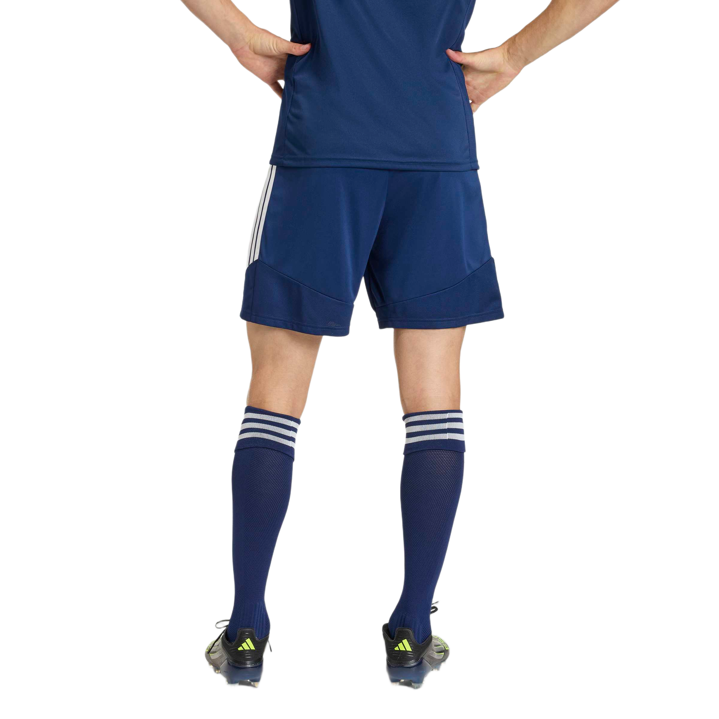 Rear view of a person in a navy blue soccer uniform and cleats, standing with hands on hips against a light gray background.
