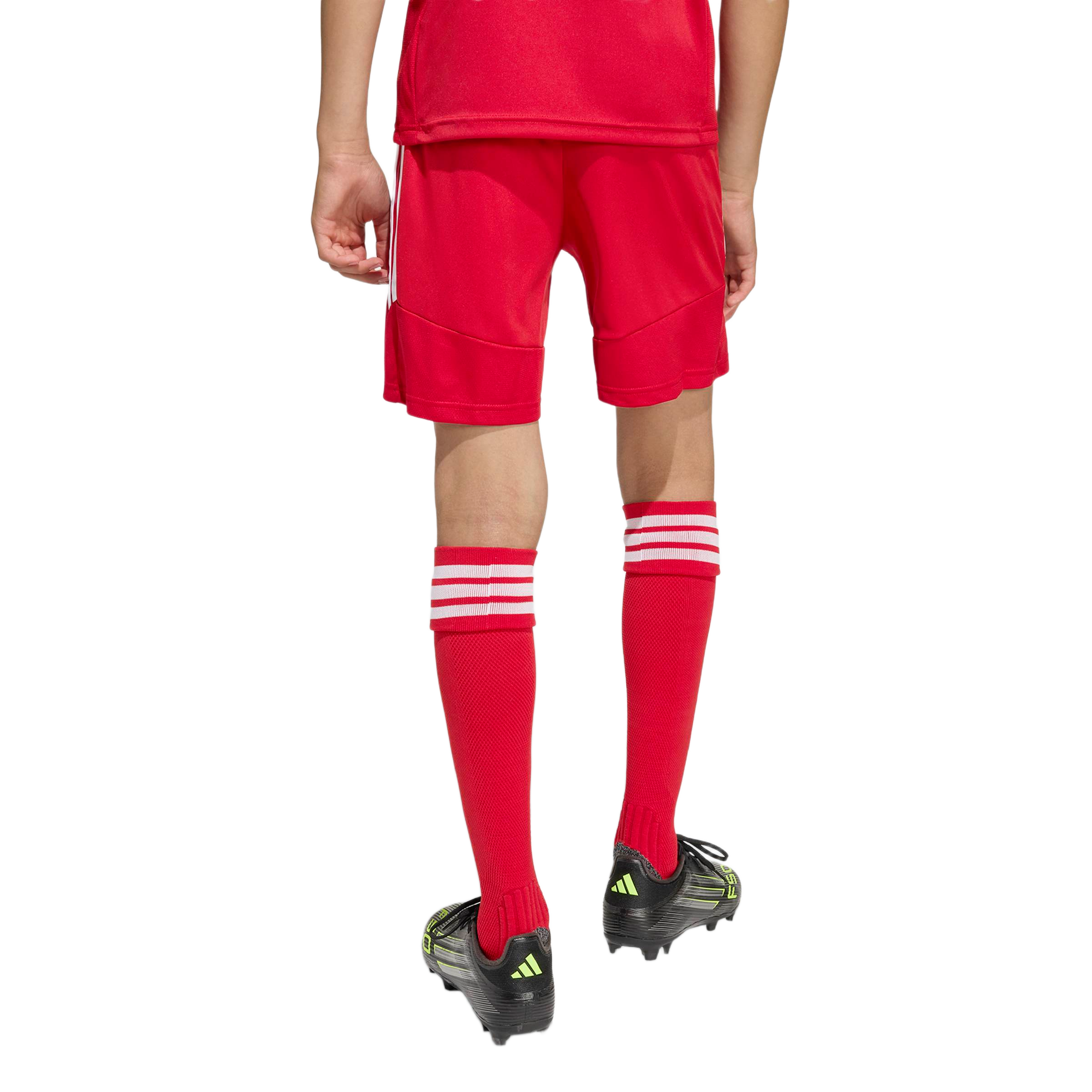 Rear view of a person wearing a red soccer uniform, including shorts and knee-high socks, and black Adidas soccer cleats, standing against a plain white background.