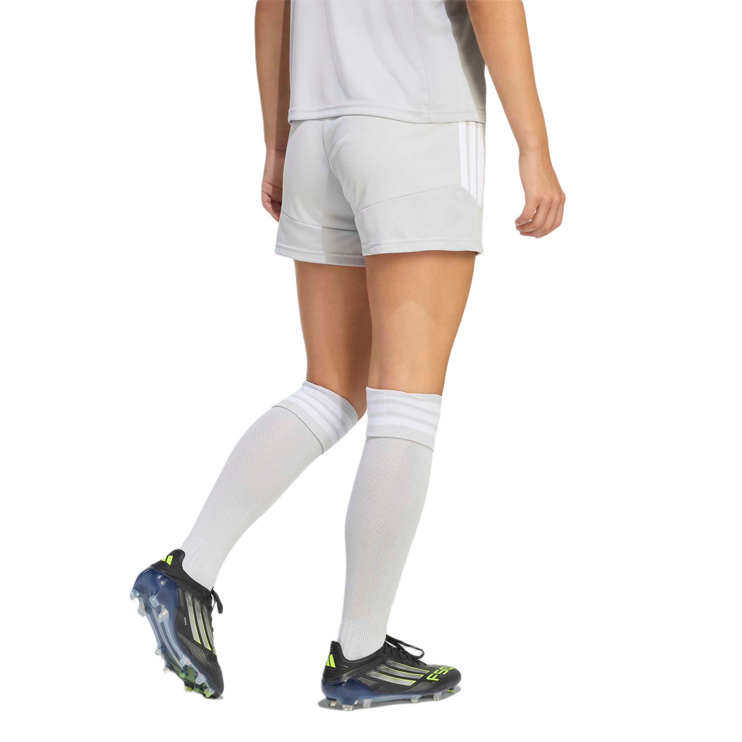 Rear view of a person wearing a light grey soccer kit, tall white socks, and black cleats with neon yellow accents against a plain background.