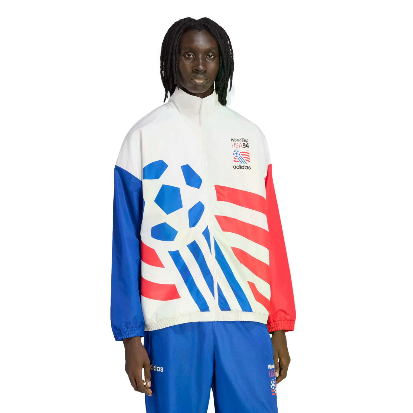 A man with dreadlocks wearing a white Adidas tracksuit jacket with red and blue graphics commemorating World Cup USA '94. The jacket features a large blue soccer ball design, red stripes, and mismatched red and blue sleeves. He is wearing coordinating blue athletic pants against a plain black background.