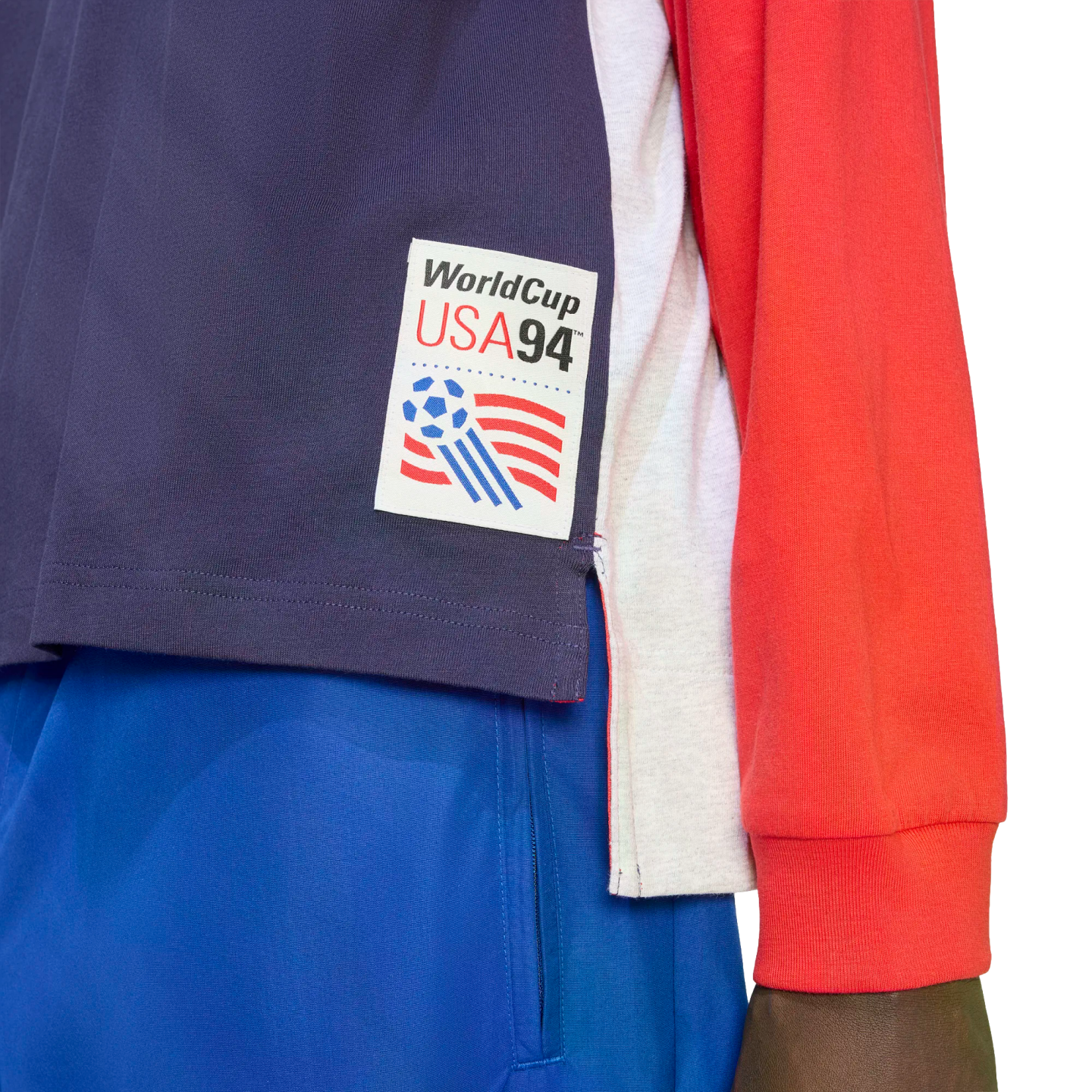 A close-up of a navy blue, white, and red athletic top featuring an official "WorldCup USA94" patch on the bottom left side. The patch includes the event's logo with a soccer ball and red stripes. The person is also wearing bright blue shorts.
