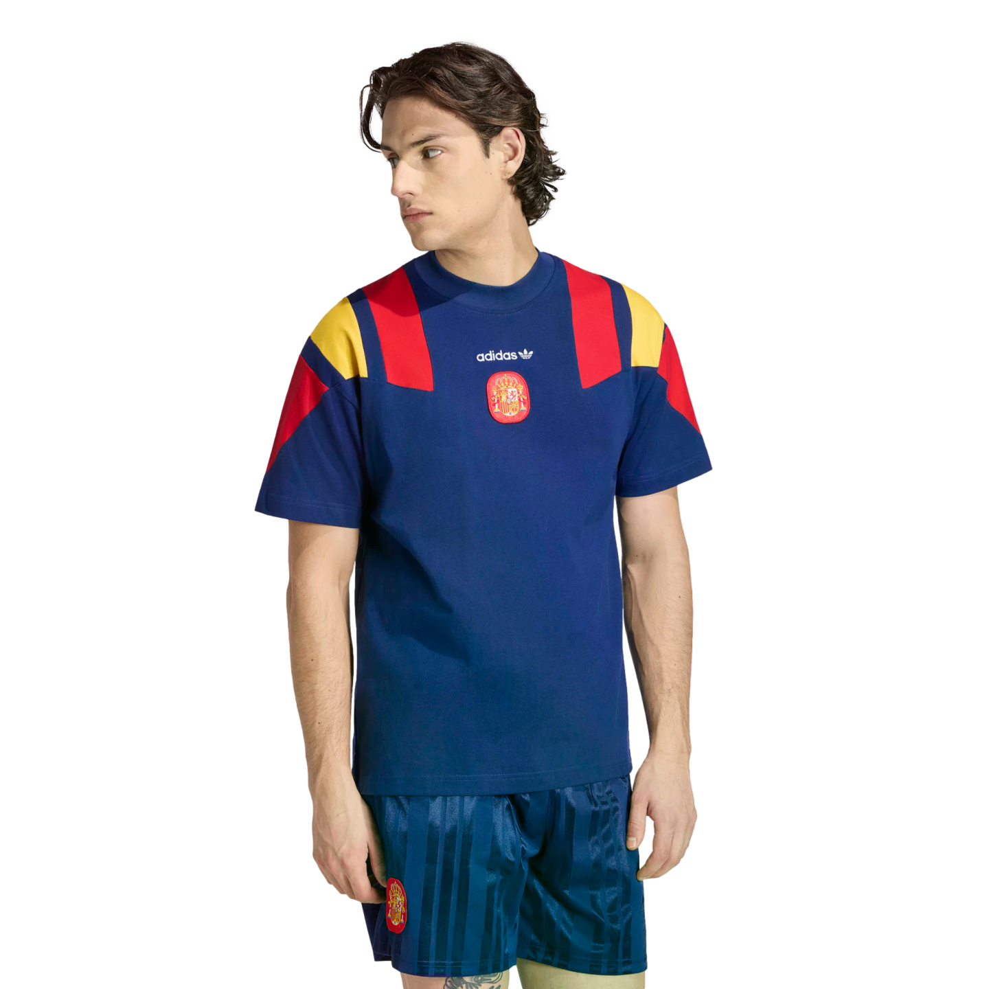 A man modeling a navy blue Adidas Spain football kit featuring a t-shirt with red and yellow shoulder panels and matching vertical-striped shorts.