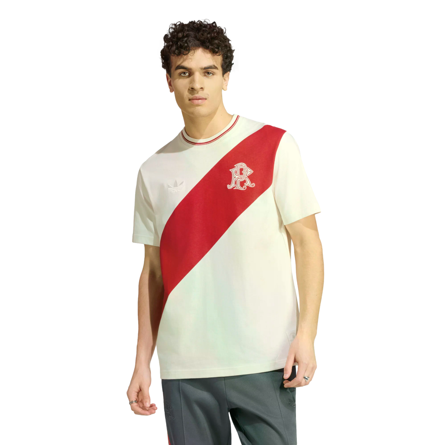 A young man with curly hair models a white Adidas t-shirt featuring a diagonal red sash and a stylized 'R' logo, paired with dark track pants.