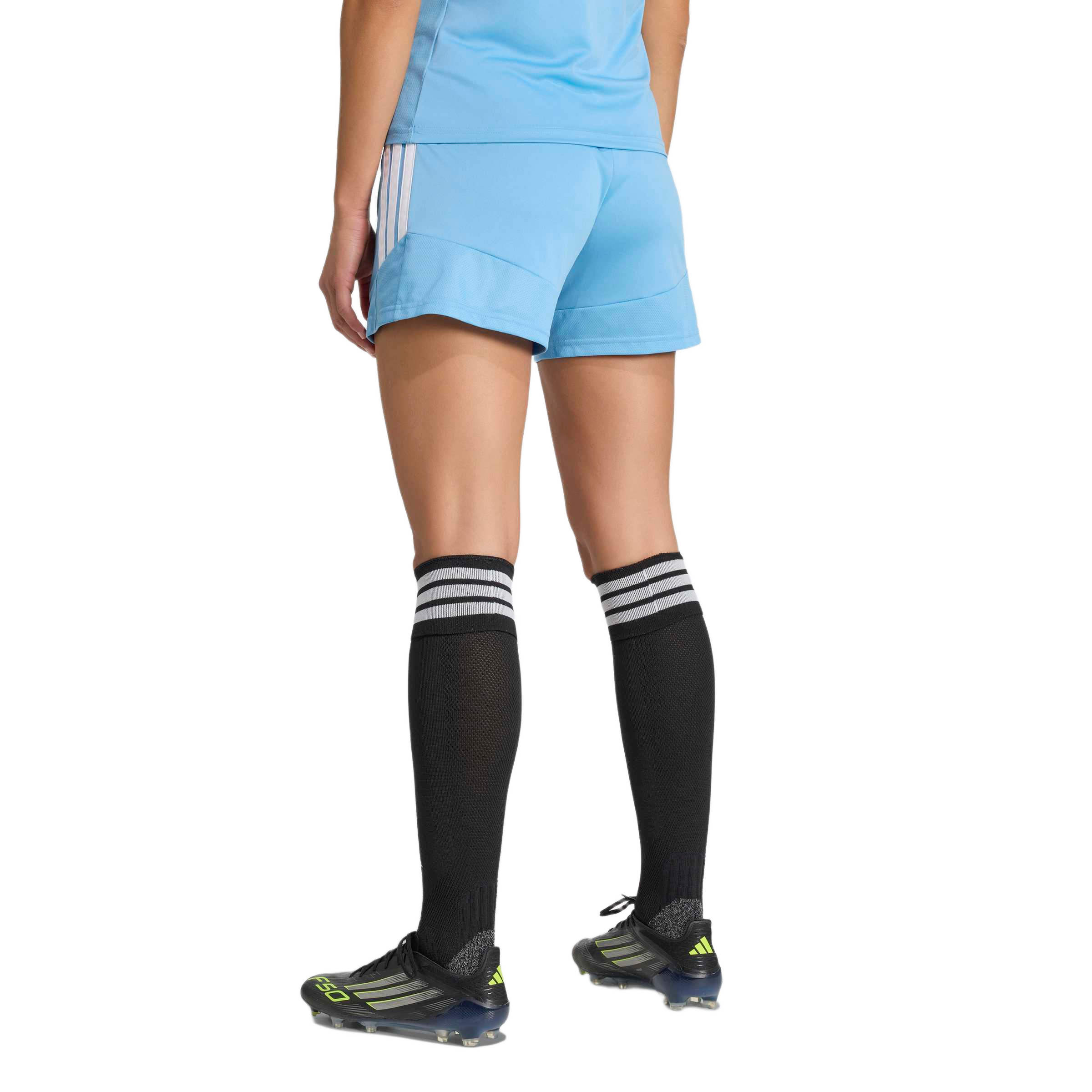 Rear view of a person wearing a light blue soccer kit, high black socks with white stripes, and black Adidas soccer cleats.