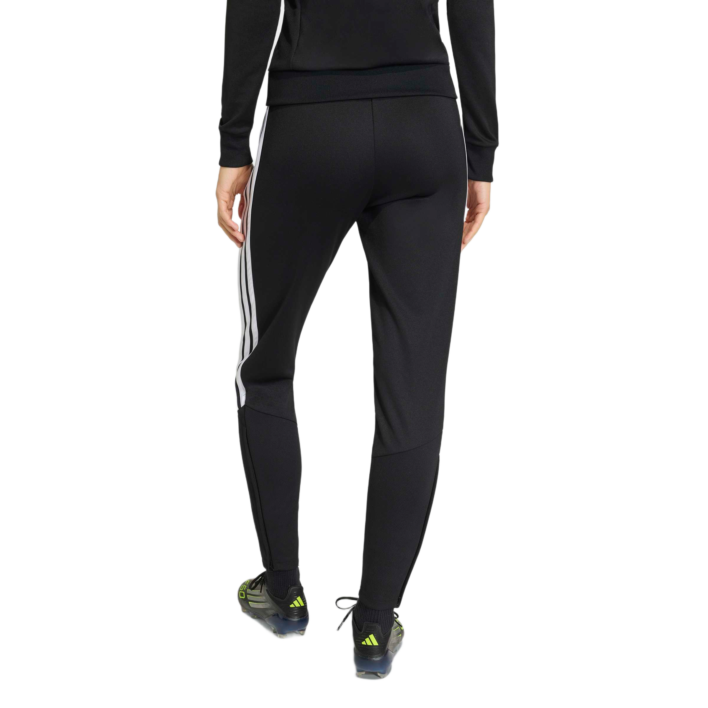 Rear view of a person wearing black Adidas track pants with white stripes on the side, a matching long-sleeved top, and soccer cleats against a light gray background.