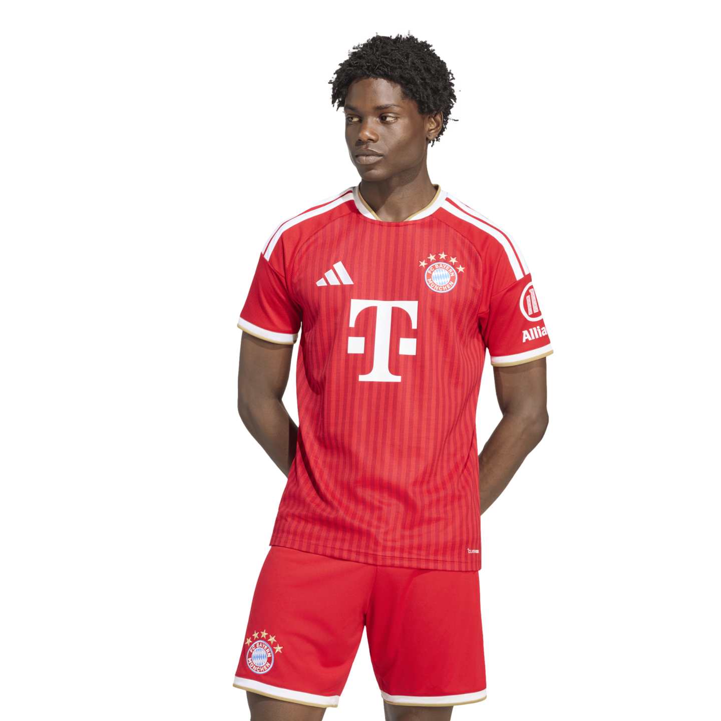 A man models a red and white FC Bayern Munich soccer kit. He stands against a white background, looking to his right with his hands behind his back.