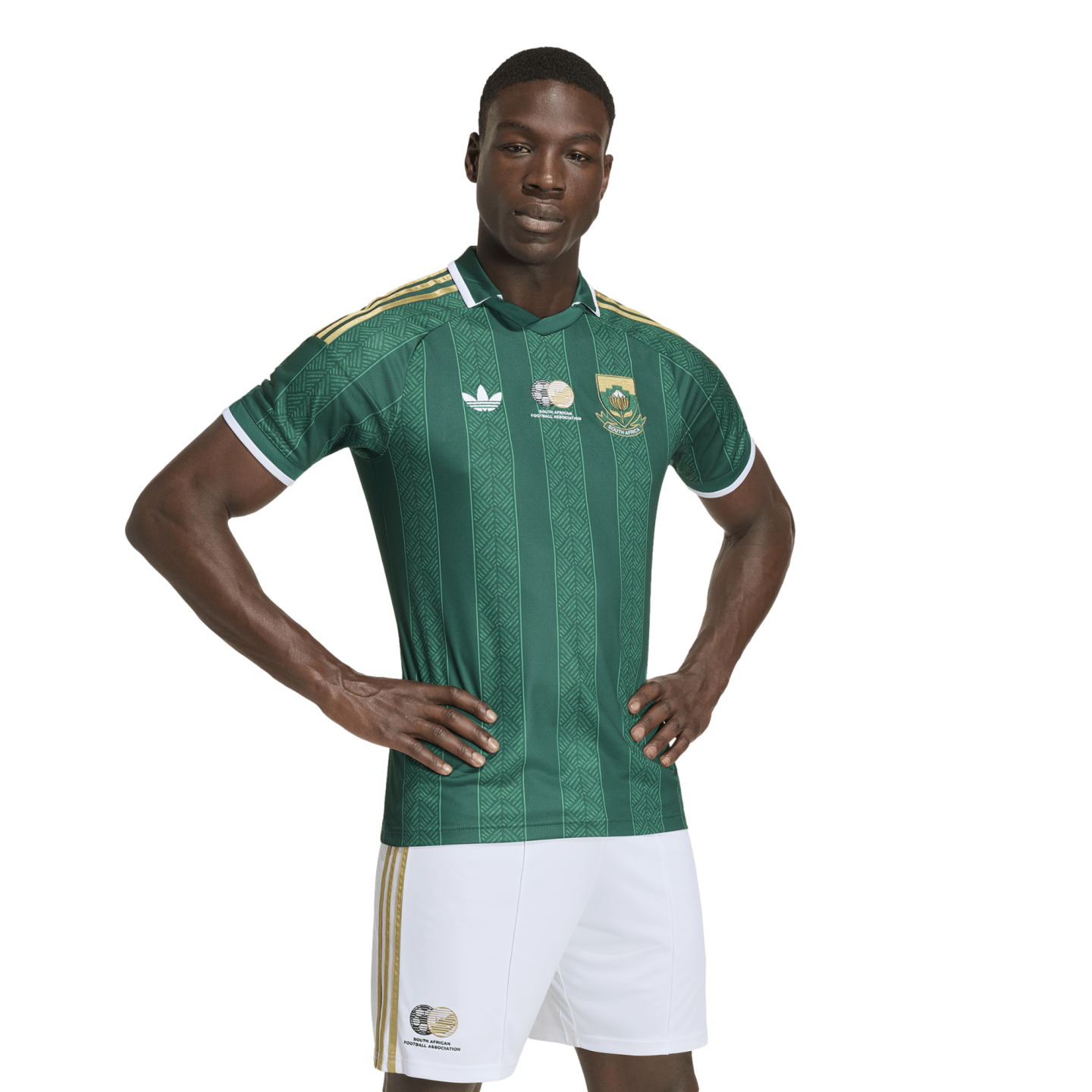 A Black man models a green and gold South African national team soccer kit, featuring a patterned jersey and white shorts, against a white background.