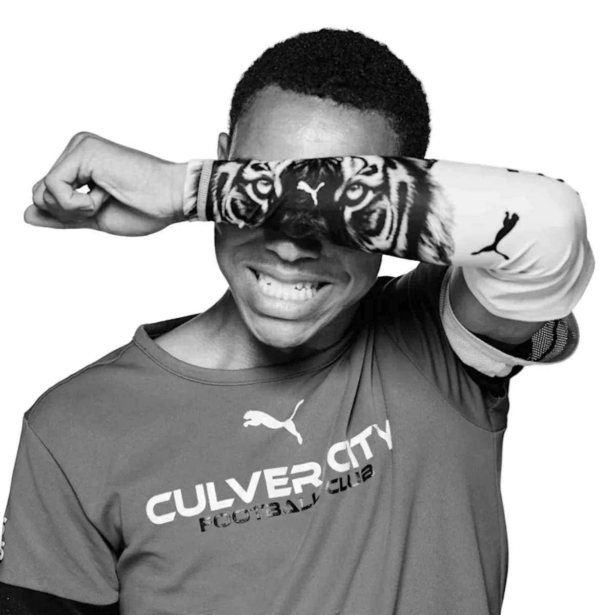 Black and white photo of a smiling man in a Culver City Football Club shirt, holding a tiger-print arm sleeve across his eyes so the tiger’s eyes align with his own.