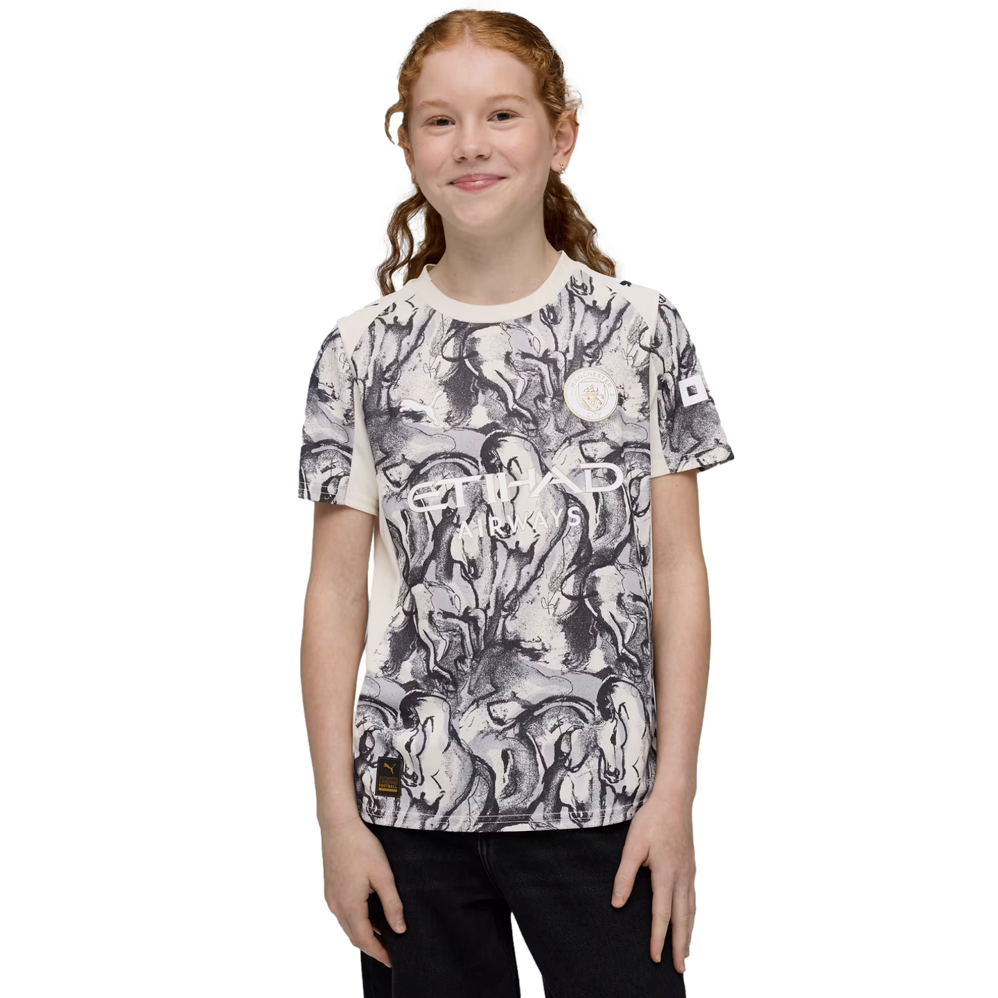 A smiling girl with red hair wears a Manchester City Etihad Airways t-shirt featuring a black and white graphic print against a black background.