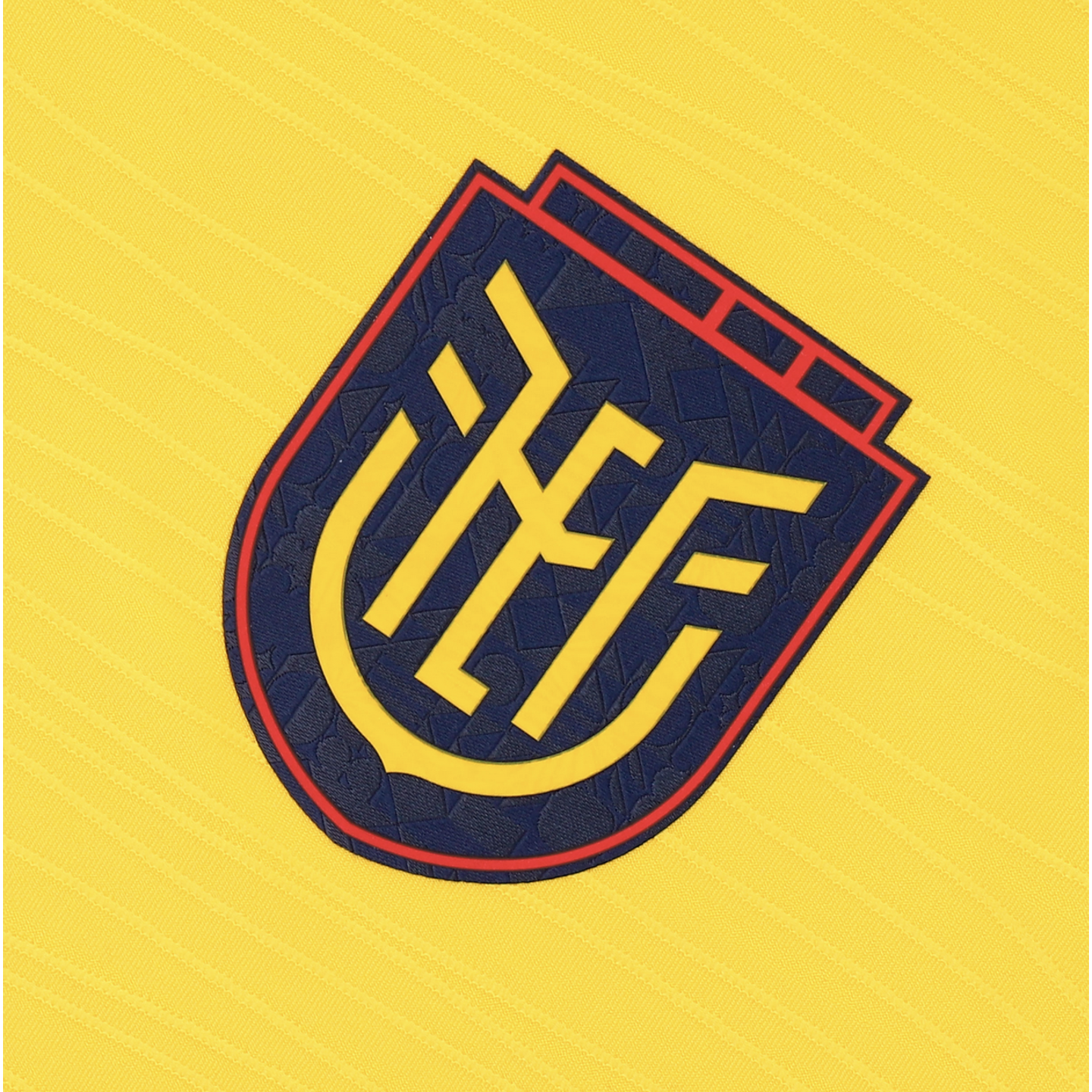 A close-up of the Ecuador national football team's crest on a bright yellow textured jersey. The dark blue, shield-shaped emblem features a red border and a stylized yellow geometric monogram in the center.